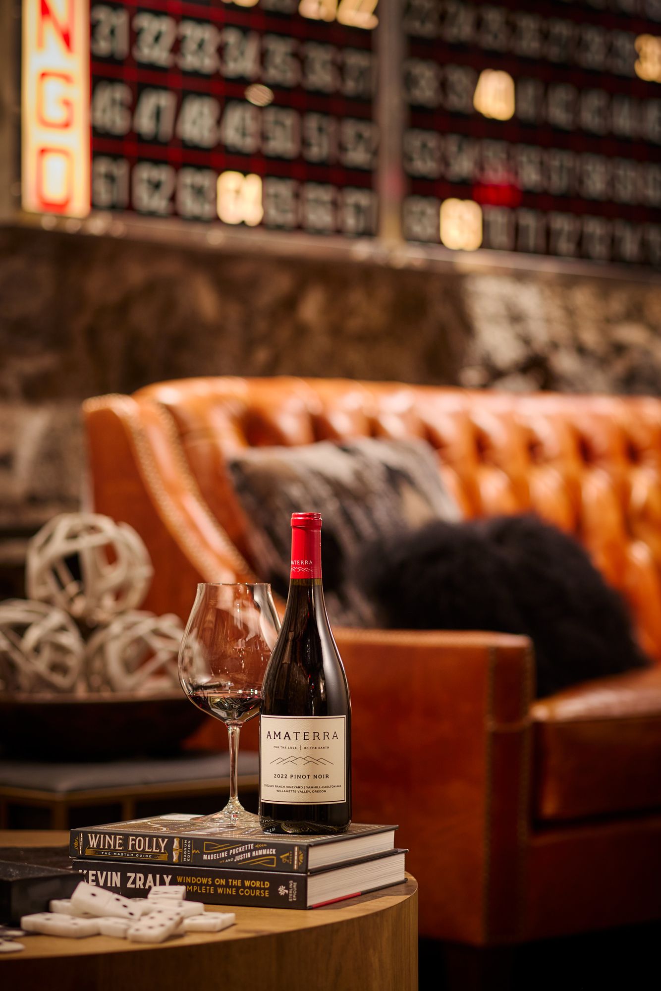 A bottle of Amaterra wine beside a glass on stacked books near a leather sofa in a cozy wine bar.