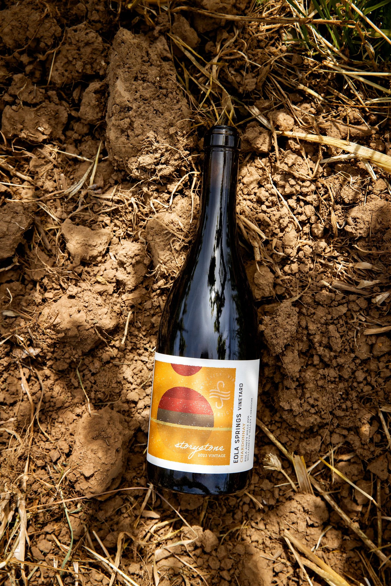Dark wine bottle with an orange and yellow sunset label lying on dry, cracked vineyard soil scattered with straw.