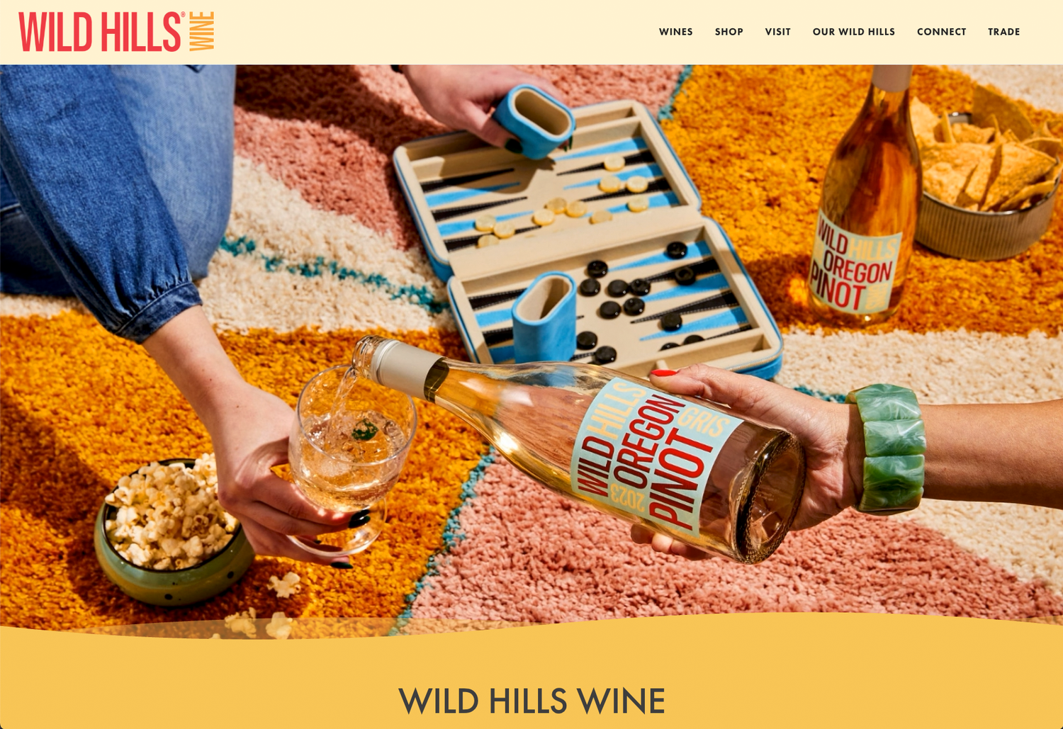 Hands pour Wild Hills Oregon Pinot Noir rosé into a glass beside a backgammon set and snacks on a colorful woven rug.