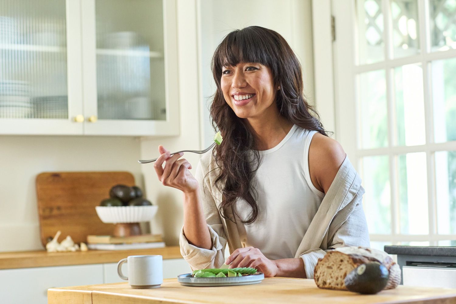 Woman sitting at kitchen counter enjoying breakfast while working on laptop in bright modern home interior.