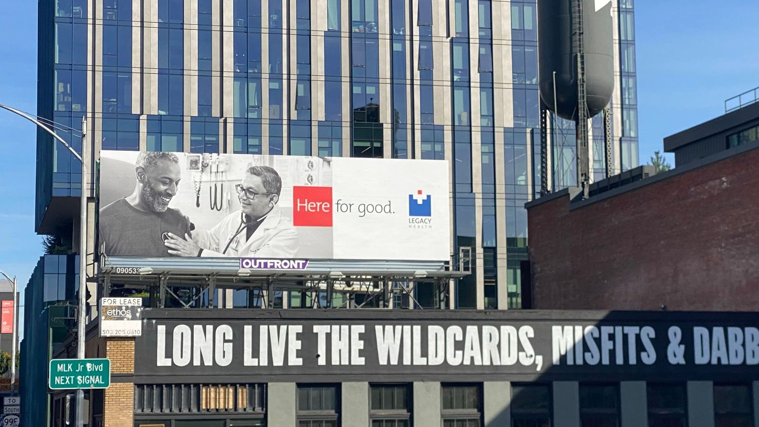Large outdoor billboard showing two smiling people with a Here for good slogan above a marquee reading Long Live the Wildcards, Misfits.