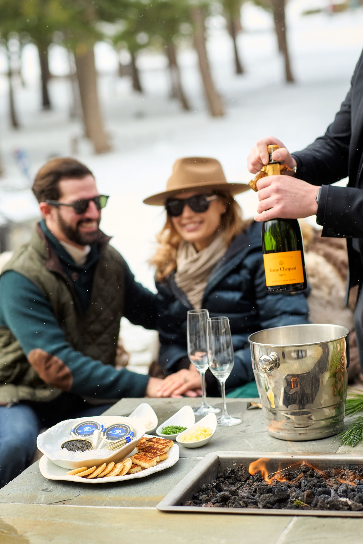 Couple enjoying champagne and appetizers by an outdoor fire pit in a snowy setting with trees in the background.