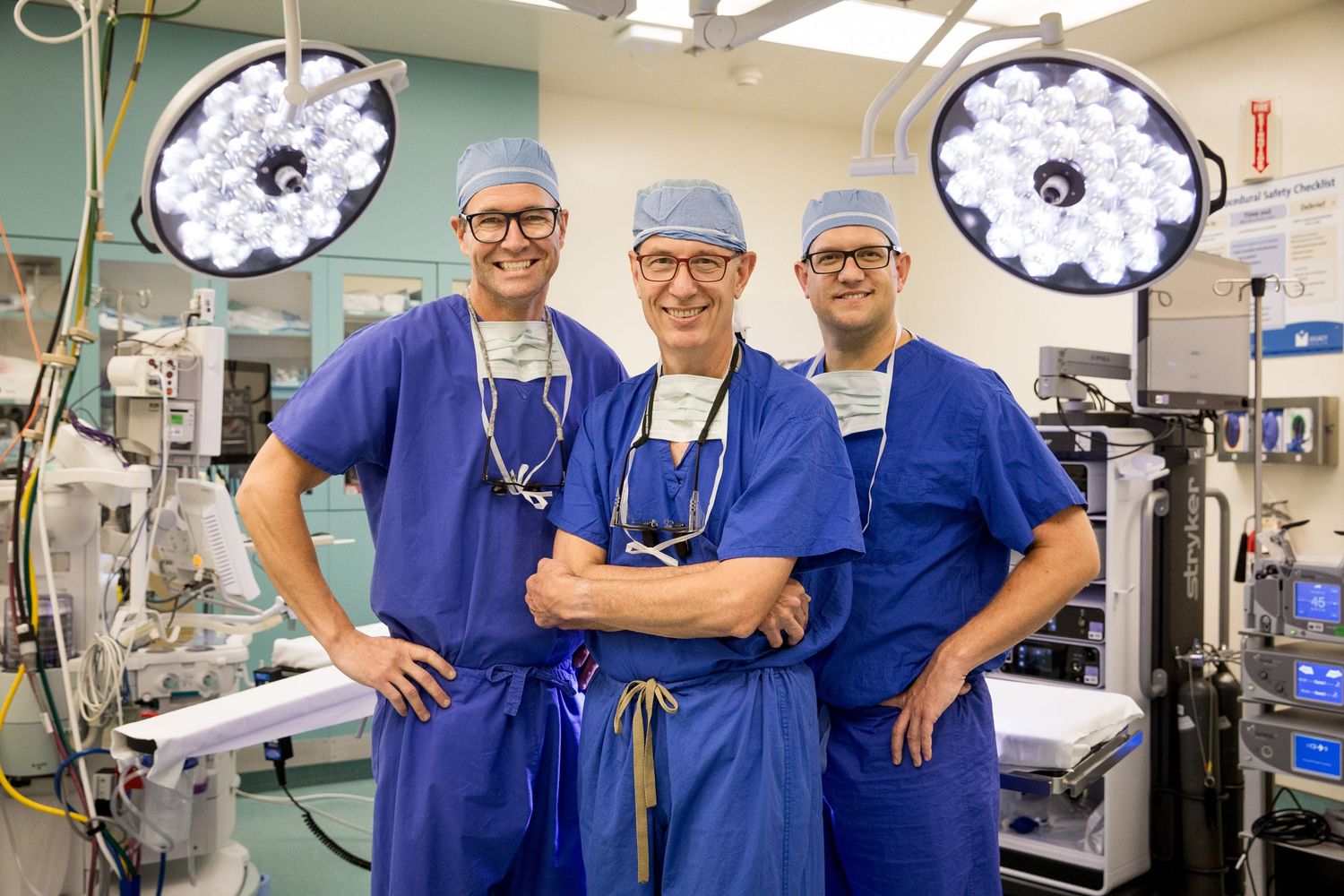 Three medical professionals in blue scrubs standing in operating room with surgical lights.