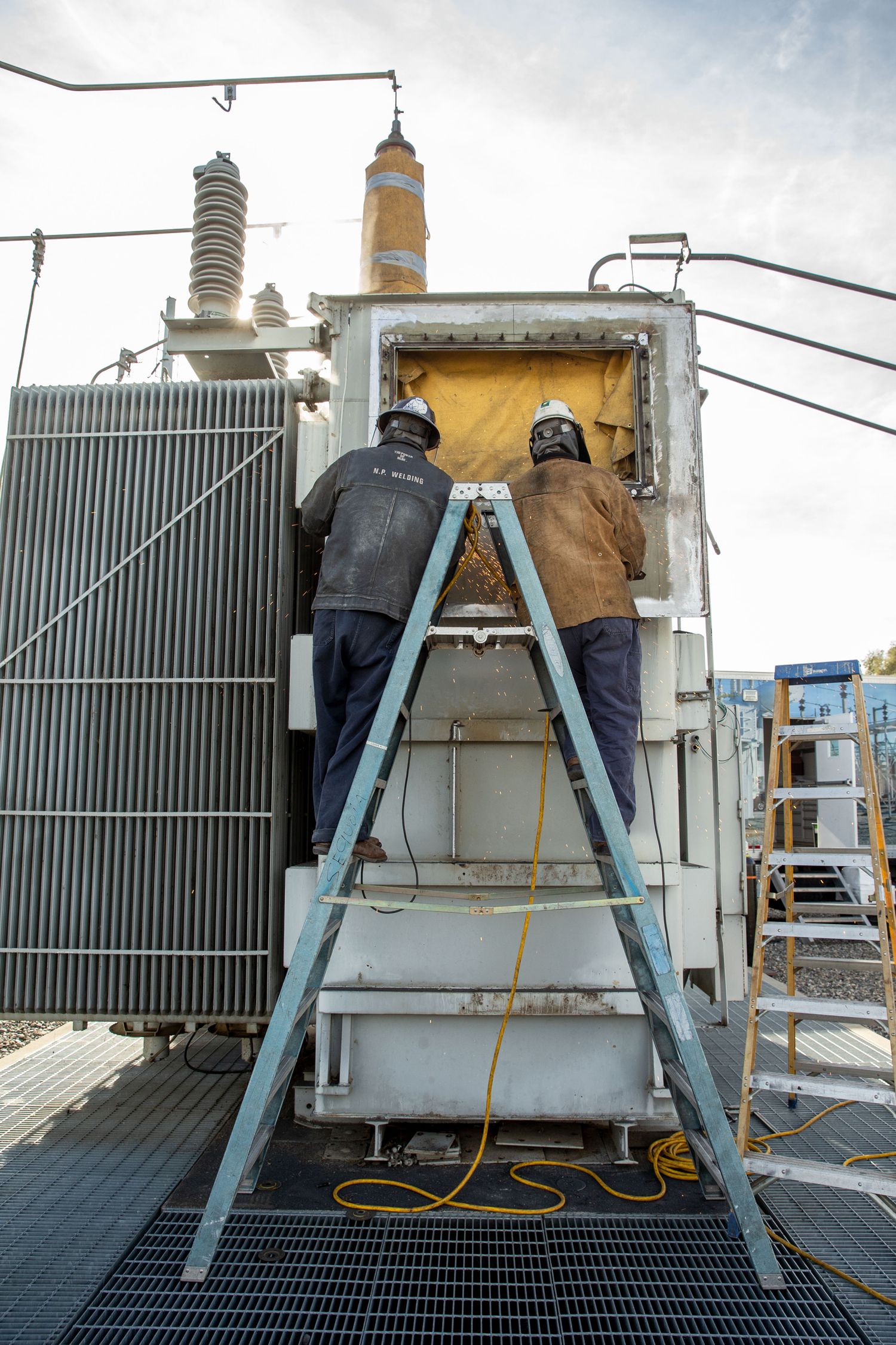 Worker on ladder performing maintenance on large electrical transformer equipment.