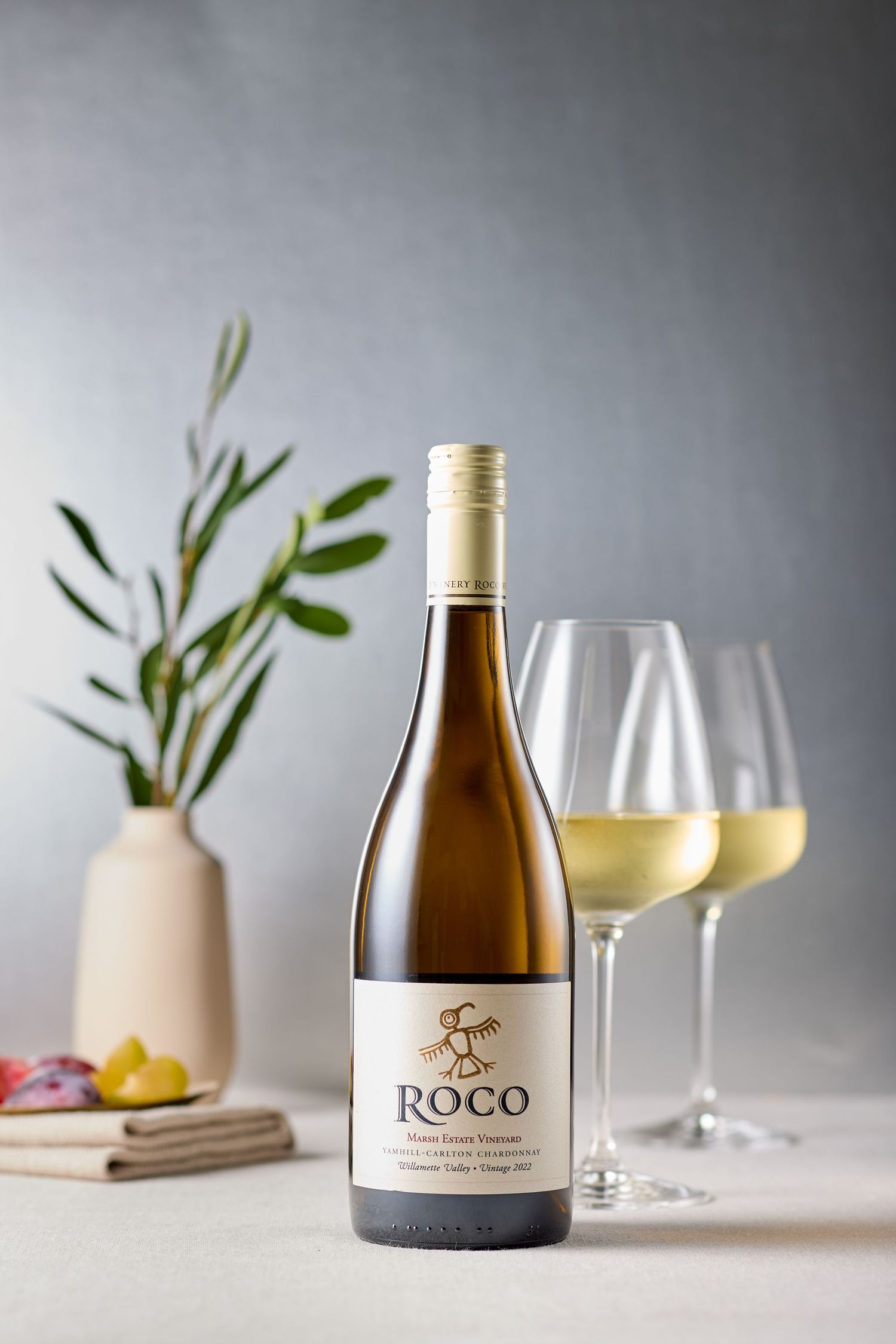 Elegant white wine bottle and filled glasses with olive branch decor on light background.