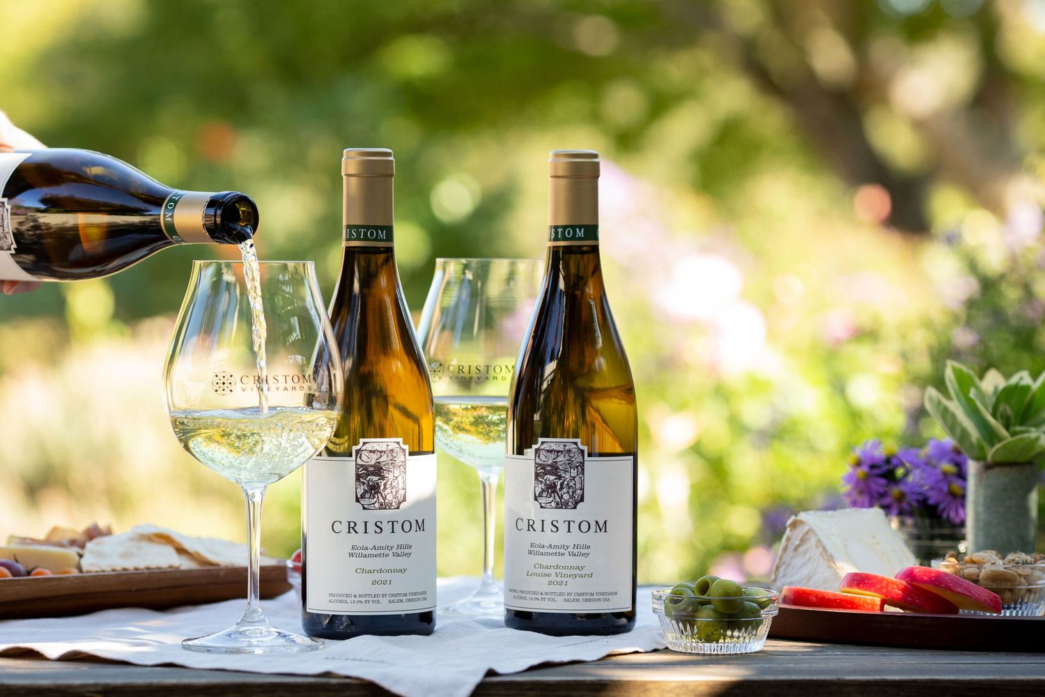 Wine bottles and glasses arranged in an outdoor garden setting with natural sunlight.