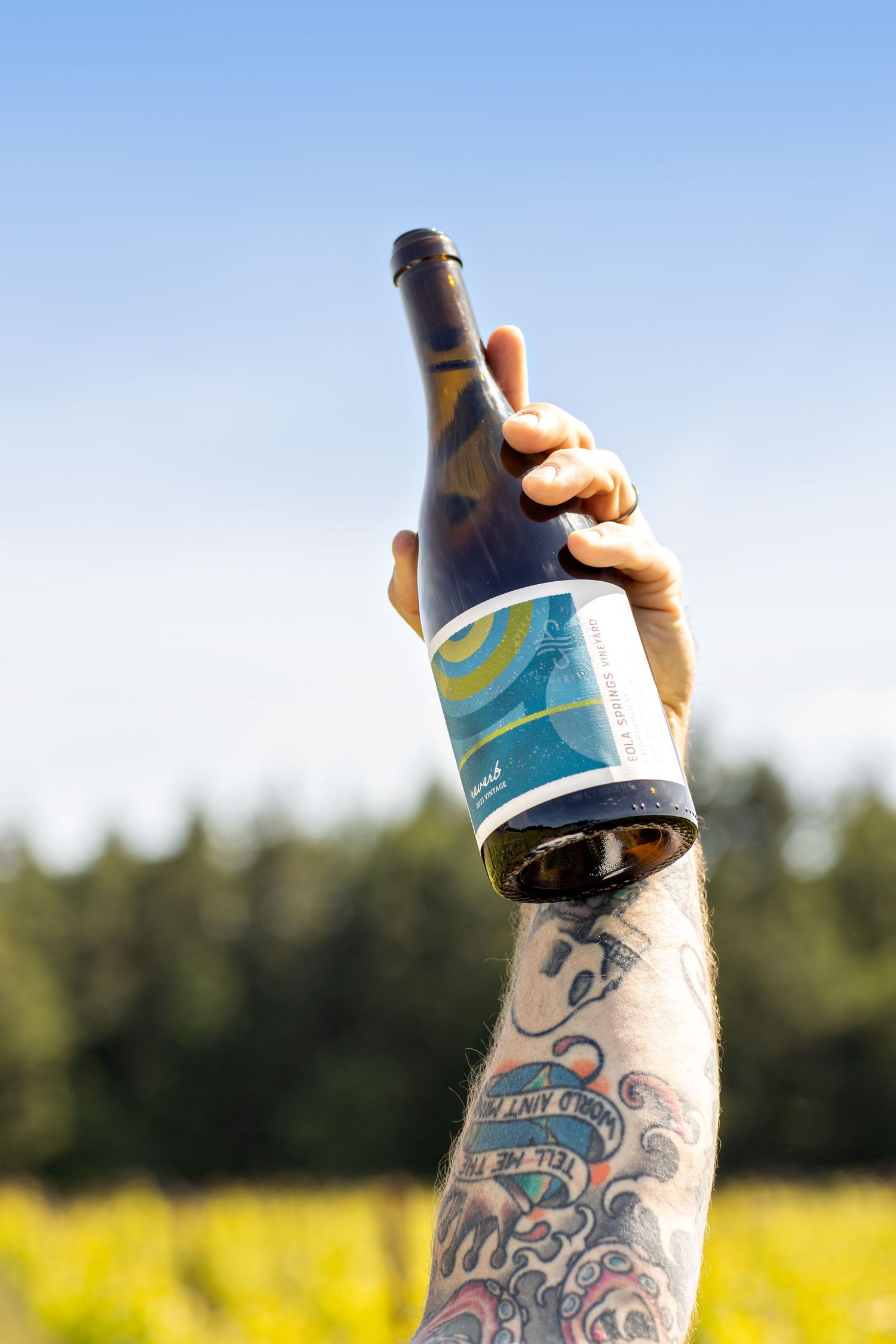 Tattooed arm holding up a wine bottle with a colorful label against a blue sky, with a yellow field and evergreen trees behind.