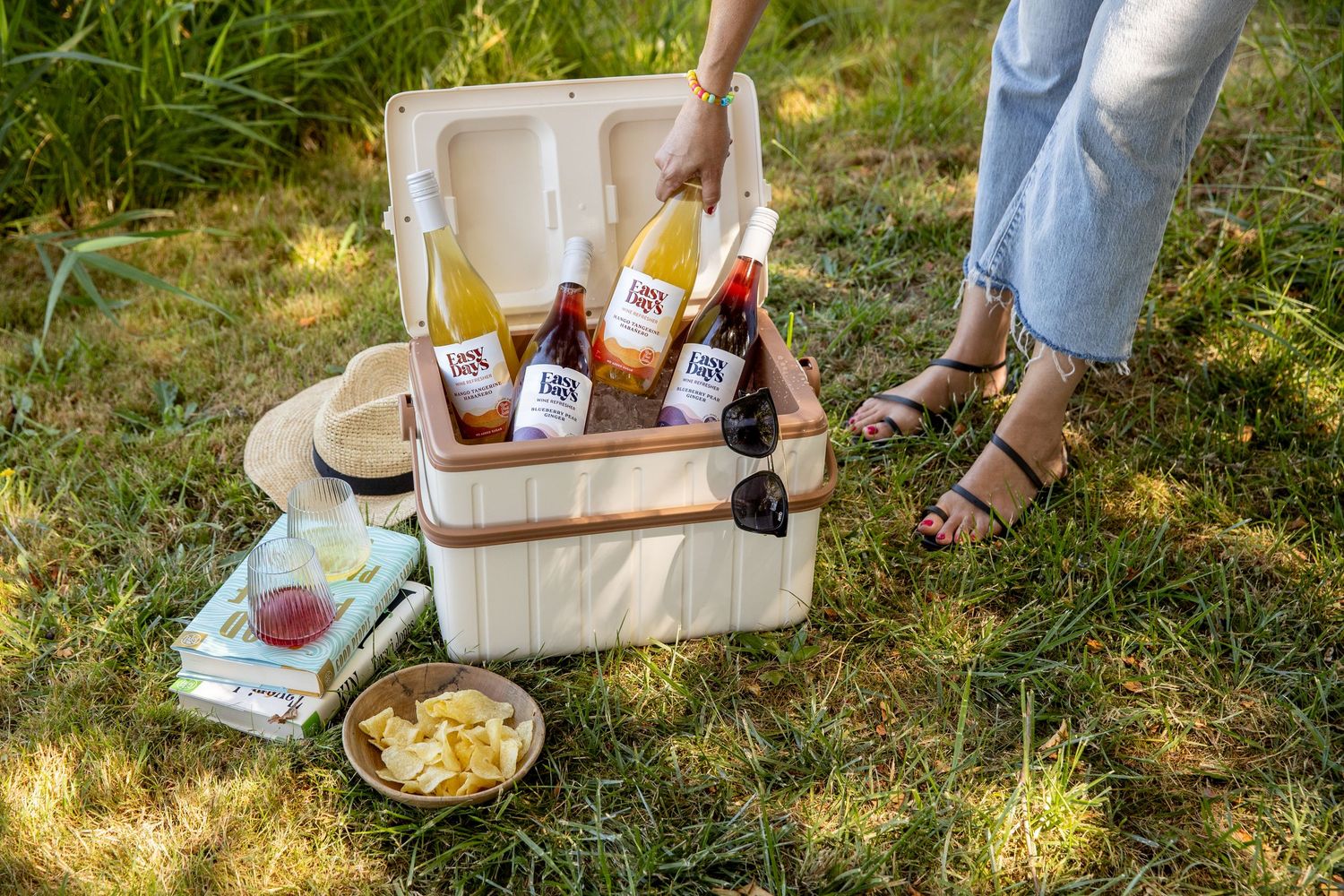 Person in jeans and sandals holds a bottle above a cream cooler of wine bottles on sunlit grass beside a snack bowl and books.