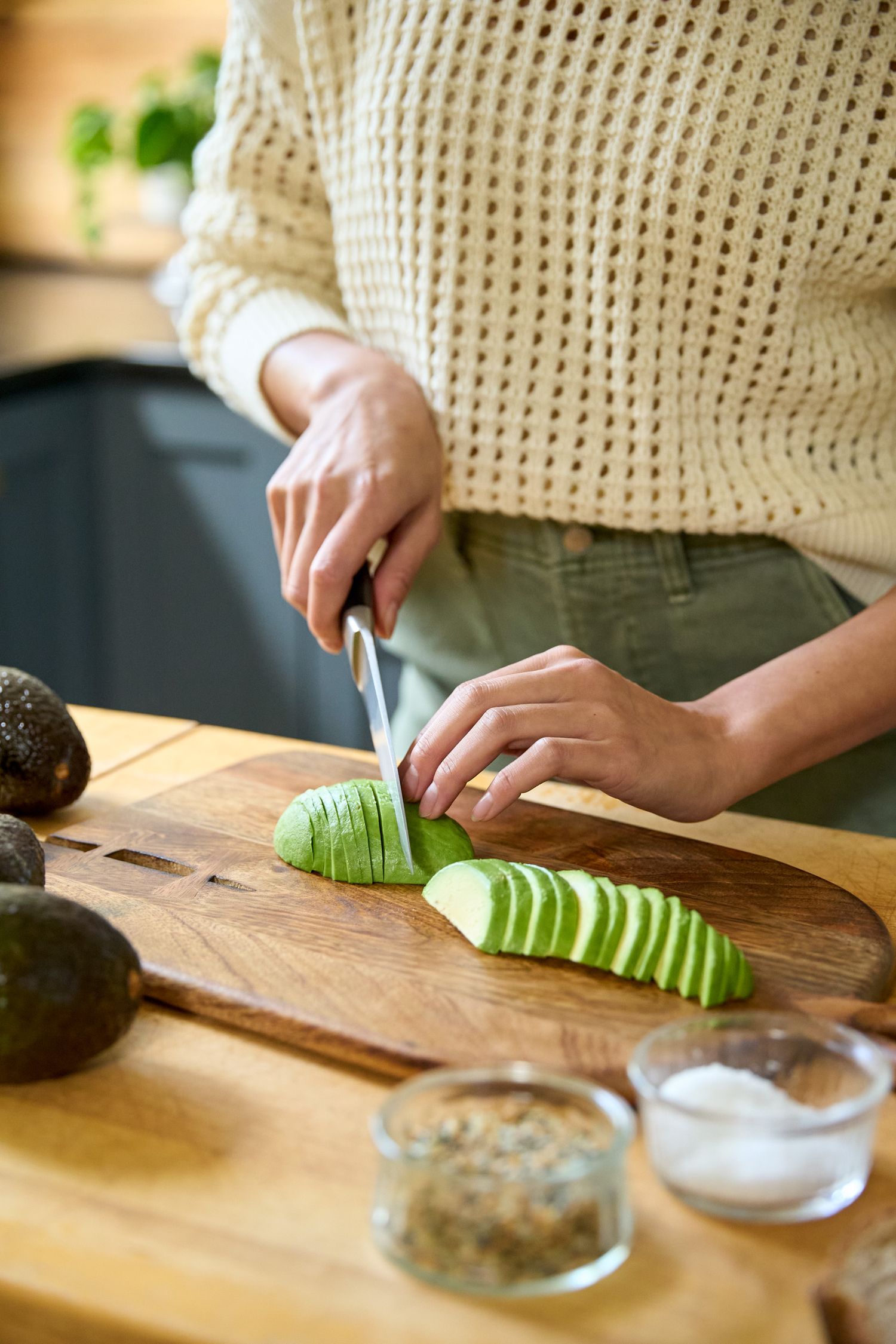 Hands slicing fresh cucumber into thin pieces on a wooden cutting board next to small glass bowls.