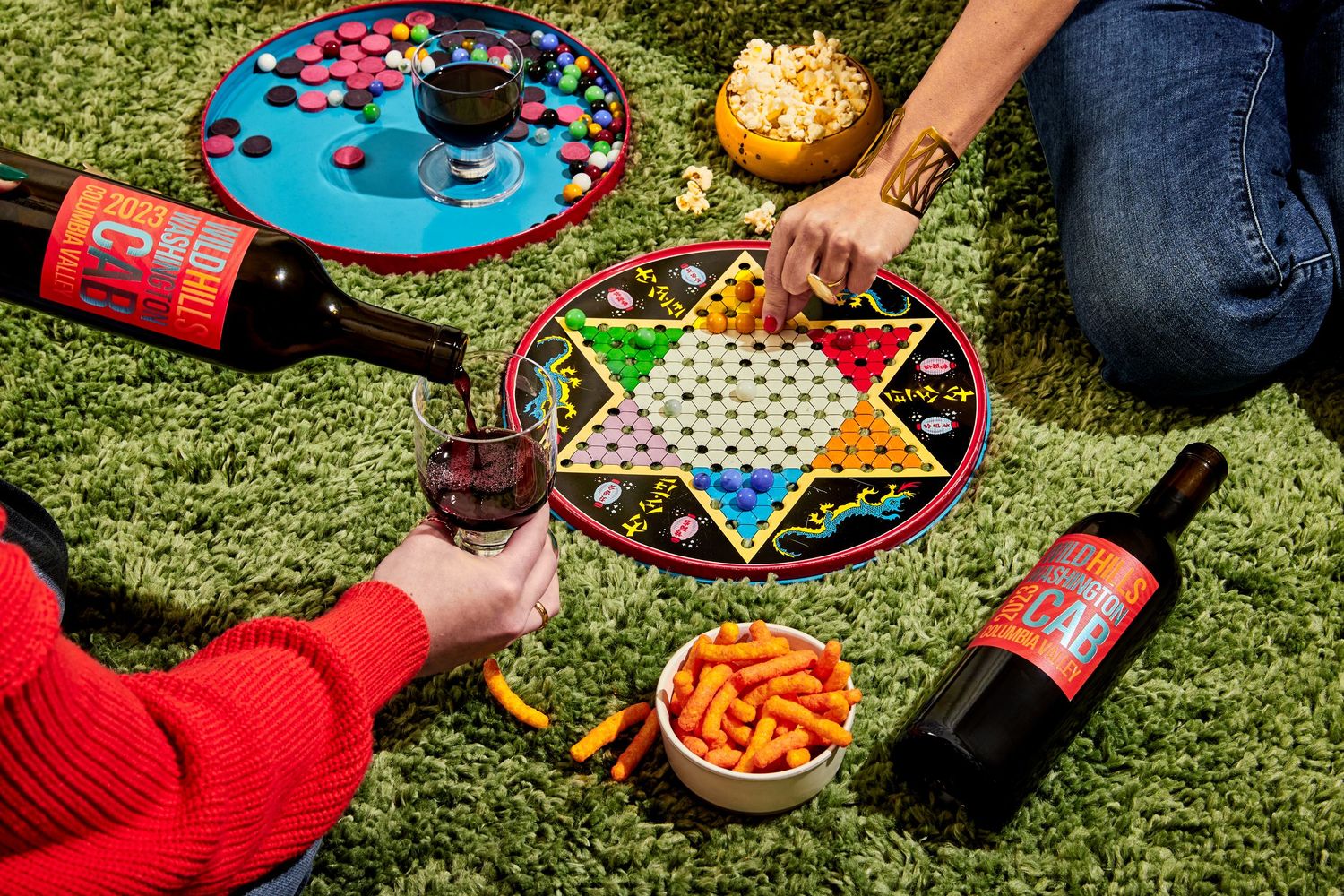 Two people play Chinese checkers on green grass with bottles of red wine, wine glasses, and bowls of cheese puffs and snacks.