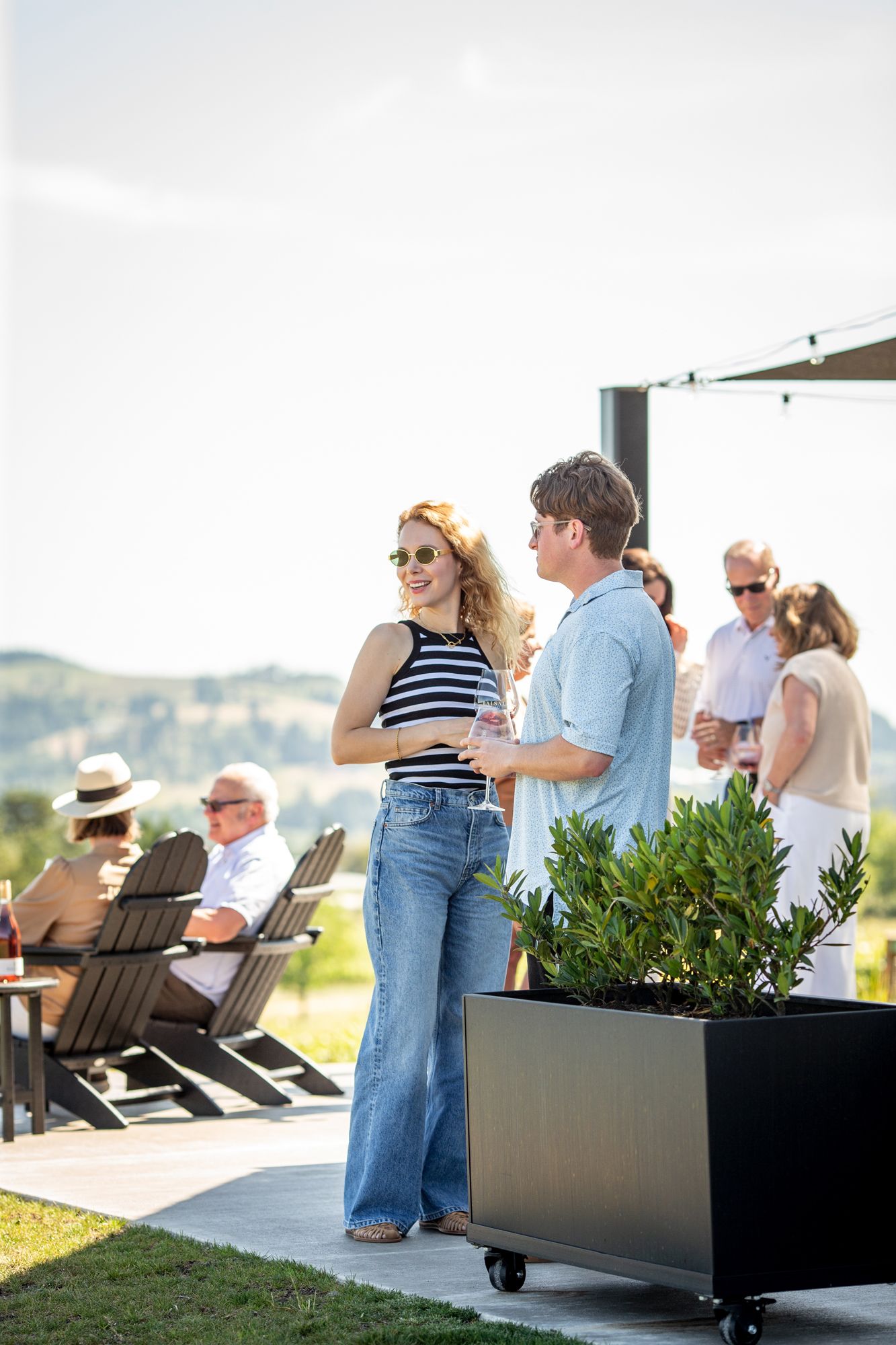 Couple socializing on an outdoor patio at a winery, with rolling hills and guests relaxing in Adirondack chairs.