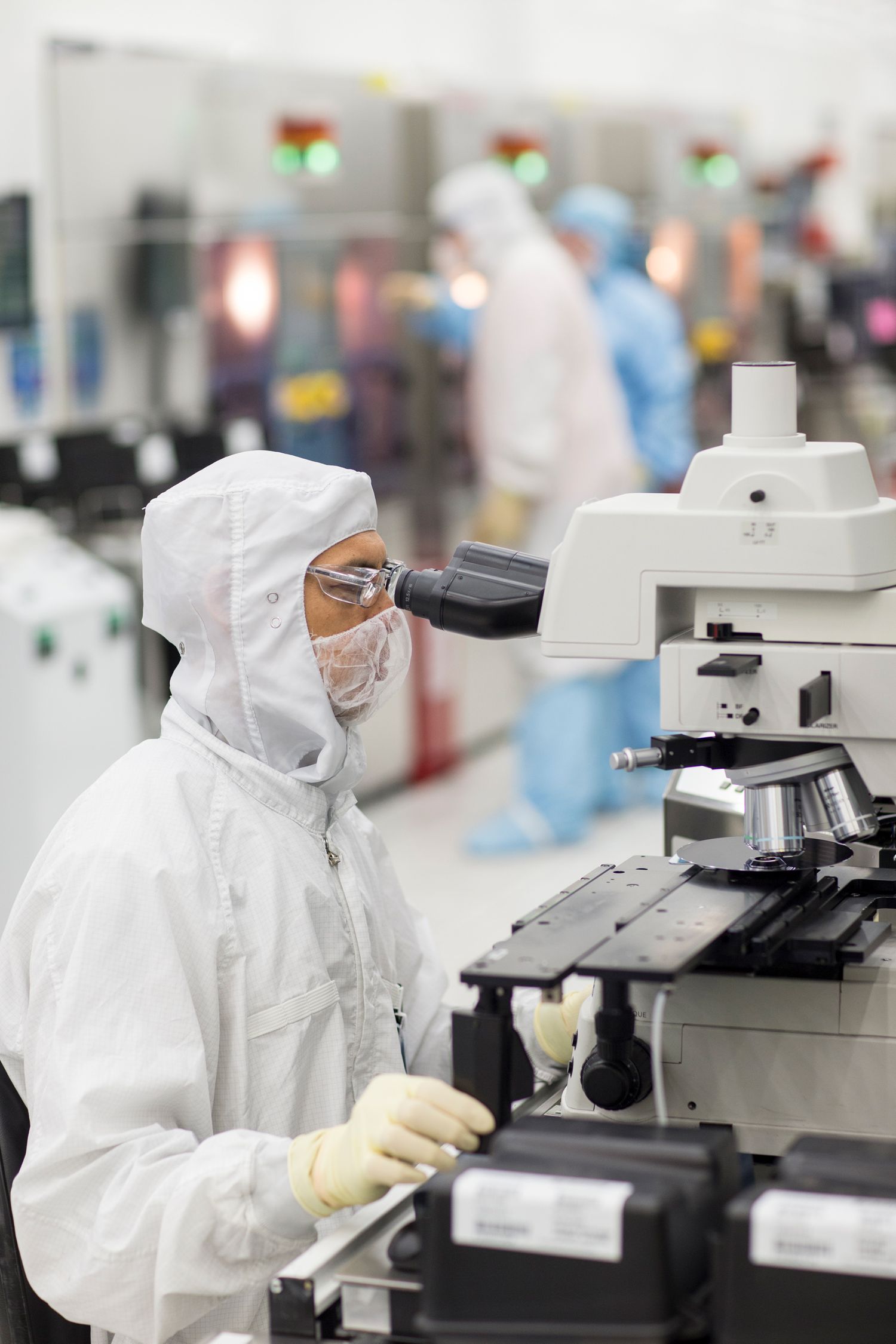 Technician in cleanroom suit examines sample through microscope in sterile laboratory.