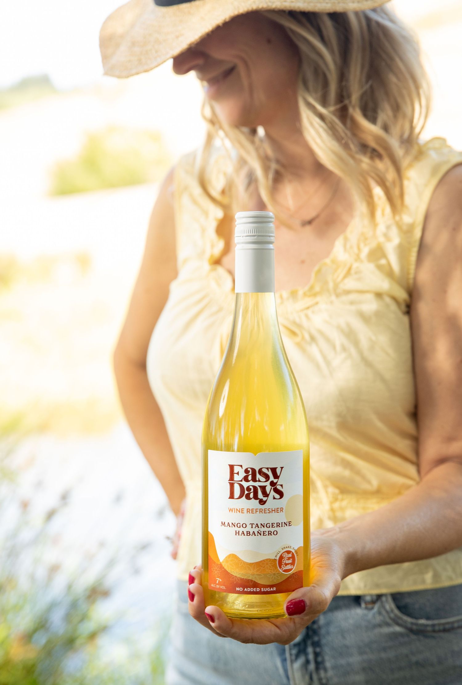 Woman in a straw hat and yellow top holds a bottle of Easy Days Mango Tangerine Habanero wine refresher outdoors in sunlight.
