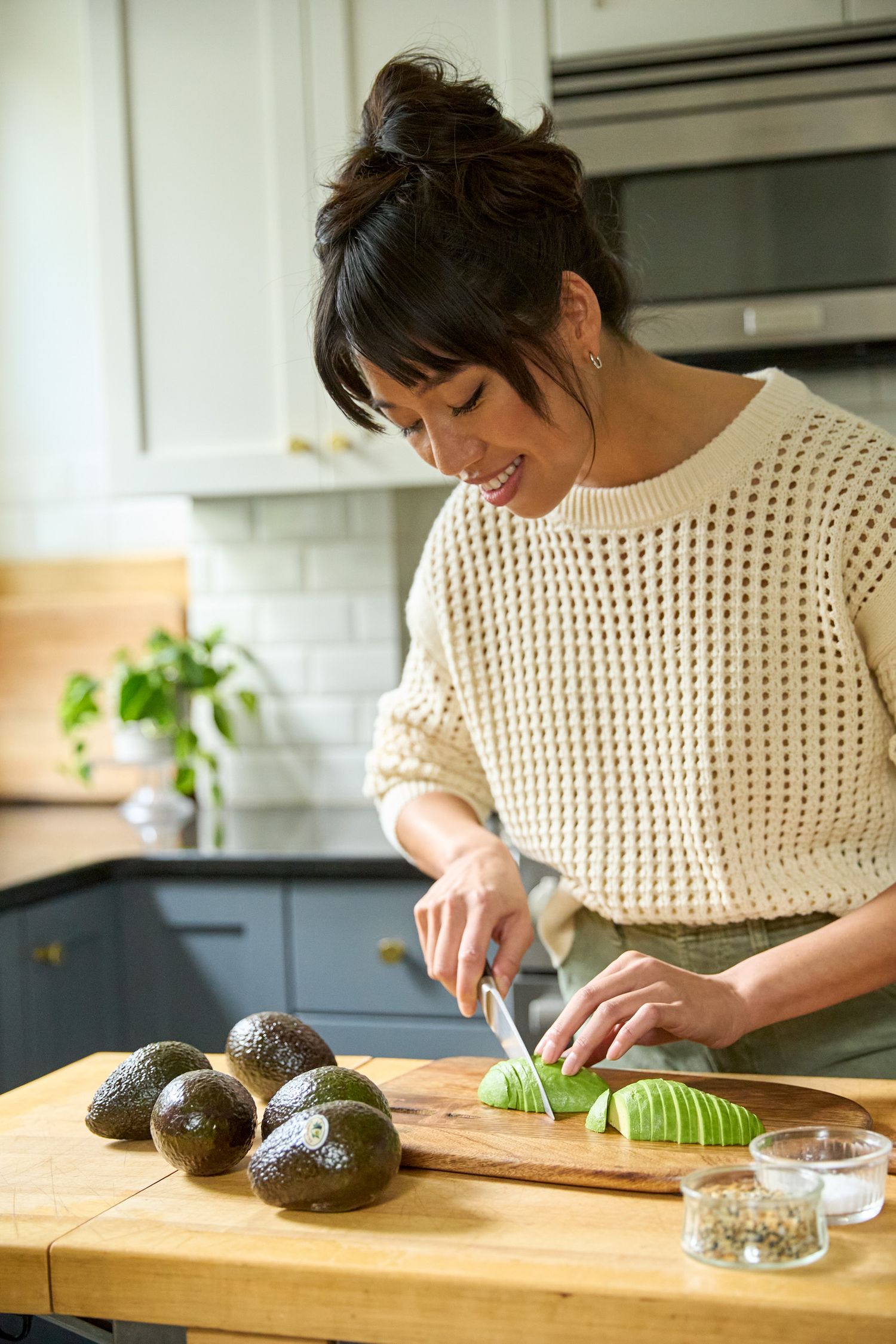 A person in a cream sweater slices fresh avocados on a wooden cutting board in a modern kitchen.