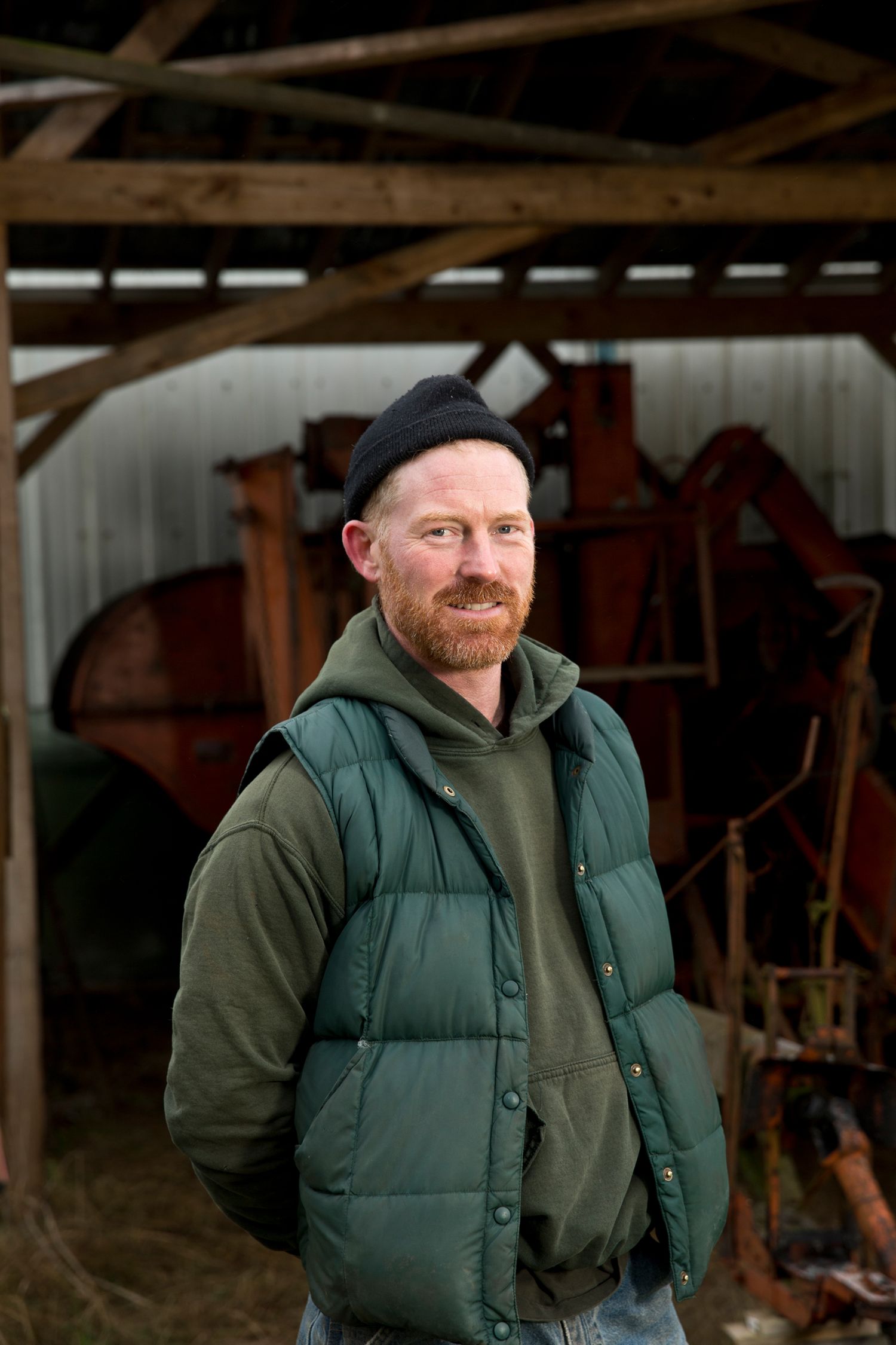 A person in a green puffy vest and beanie stands in a rustic barn with farming equipment.