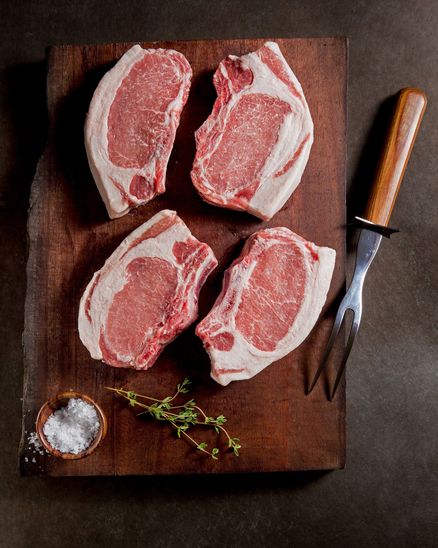 Raw thick-cut pork chops on a wooden board with herbs and salt.