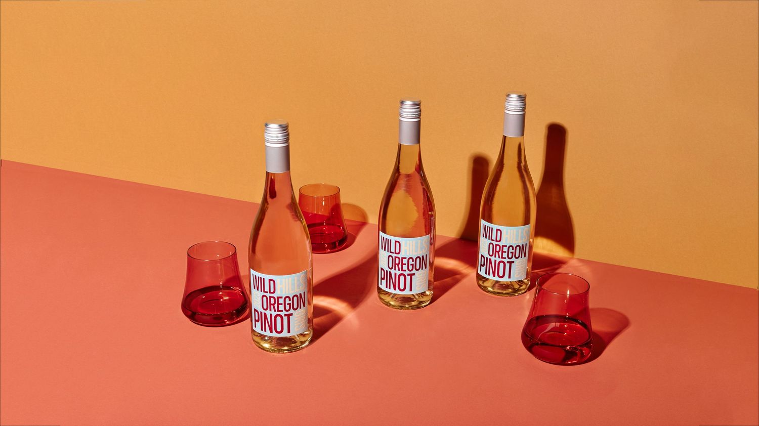 Three bottles of Wild Oregon Pinot rosé with red glasses on a coral and orange background.
