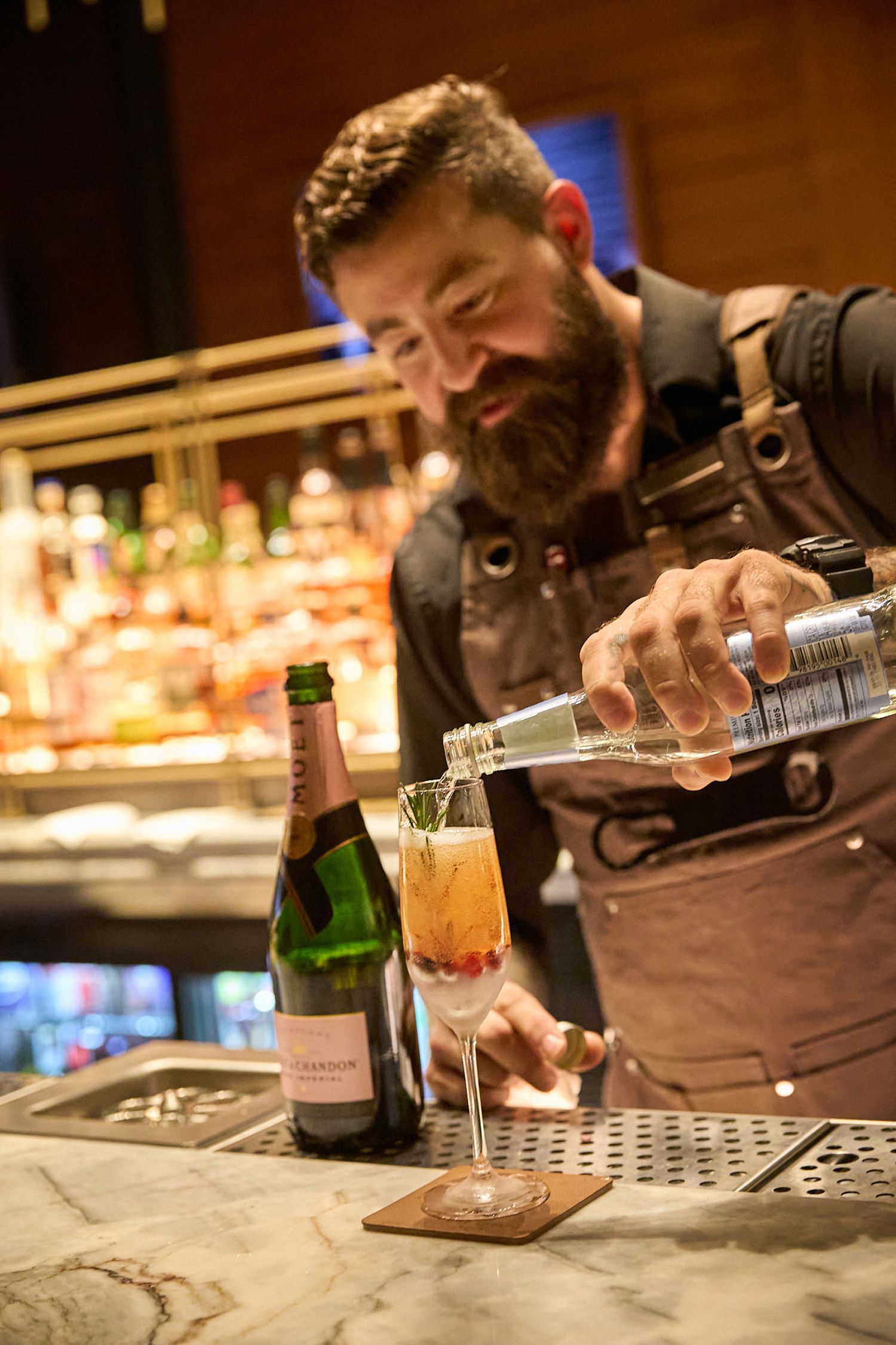 Bartender carefully pouring drinks at a dimly lit bar counter with bottles in the background.