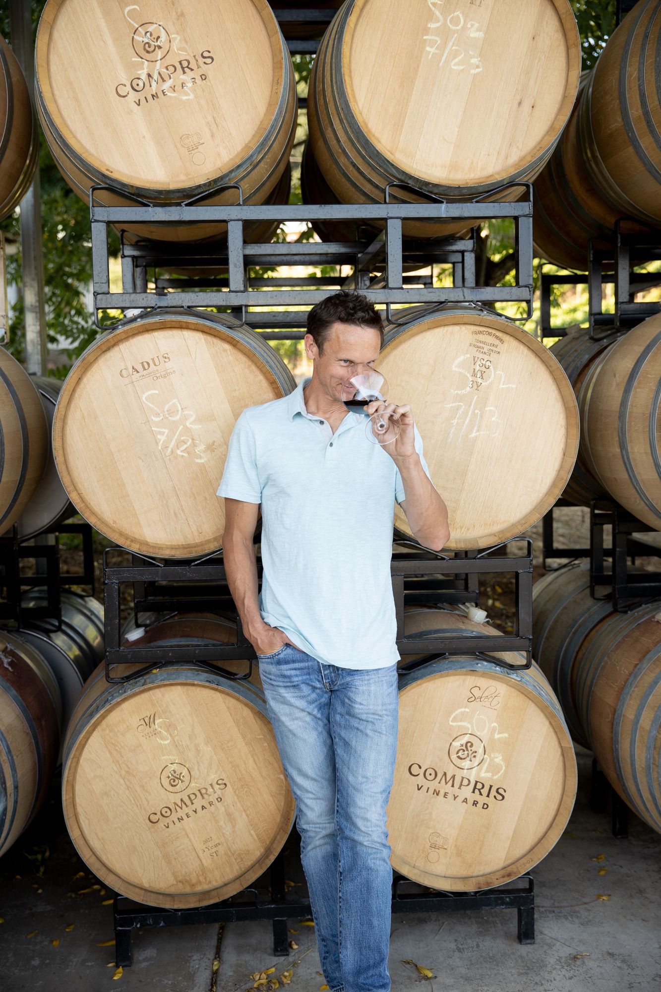 Man tasting red wine in front of stacked oak barrels at Compris Vineyard winery.