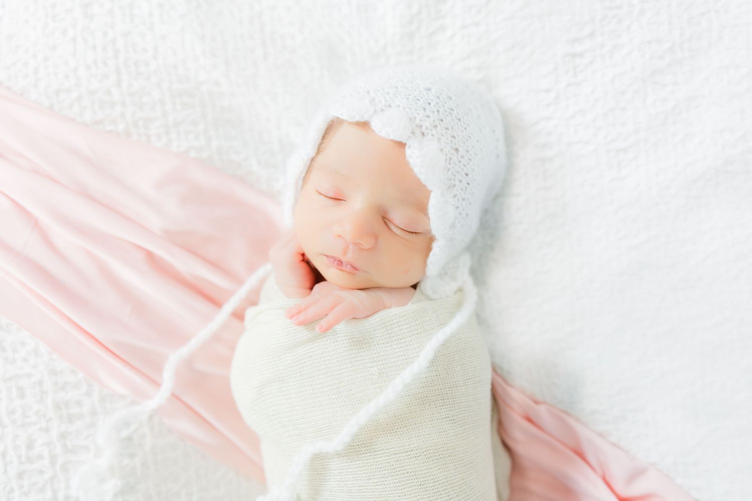 Sleeping newborn wrapped in white swaddle with matching cap on pink and white backdrop.