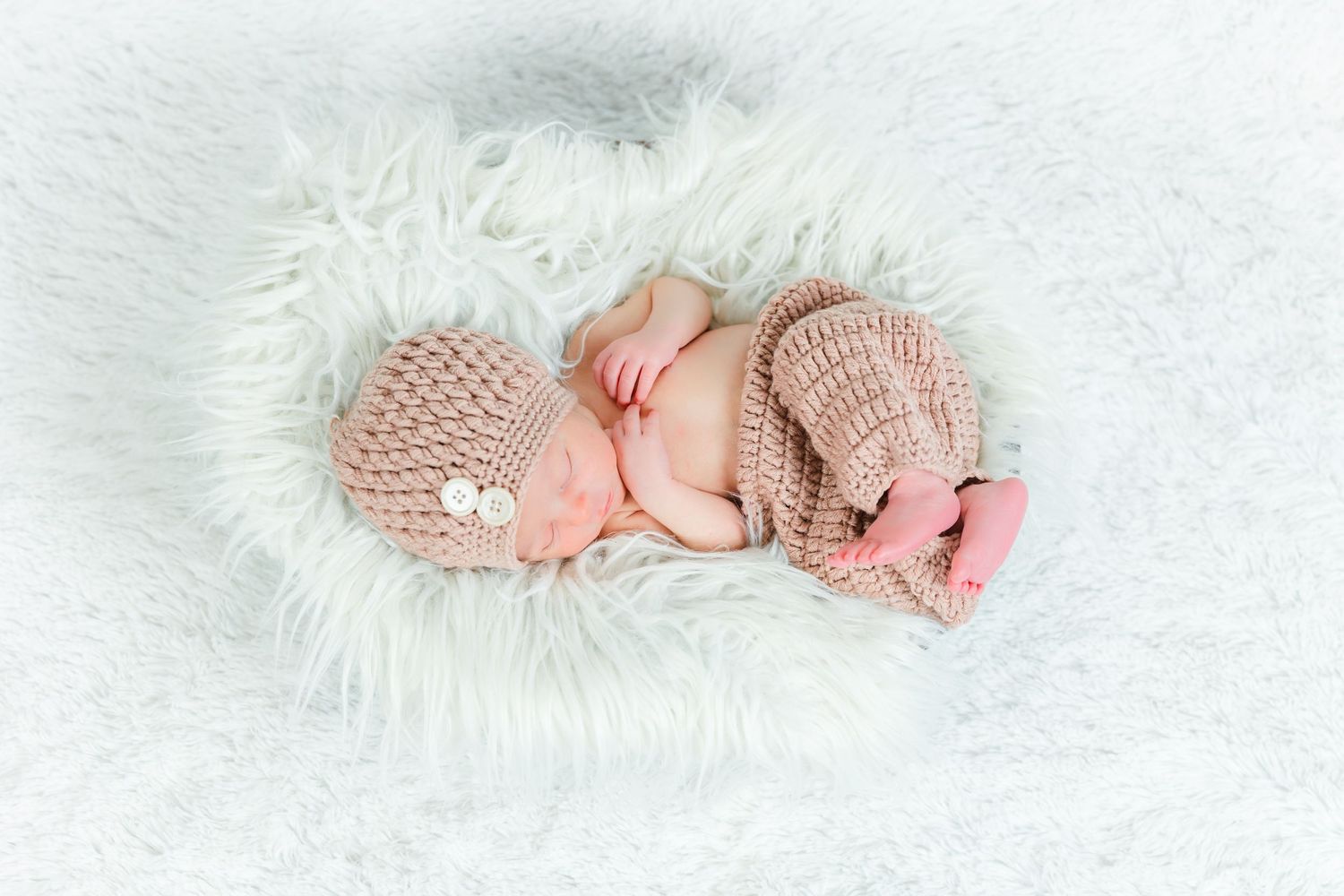 Sleeping newborn baby wearing knitted pink outfit on white fur blanket.