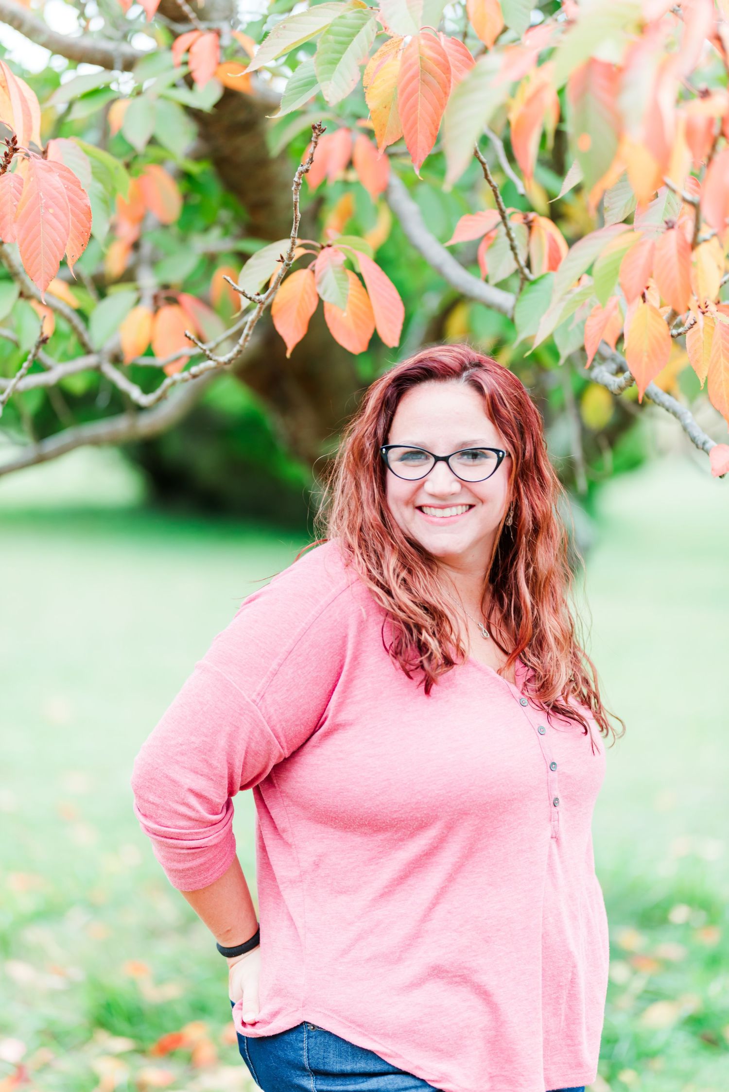 Woman in pink sweater smiles beneath autumn leaves wearing glasses.