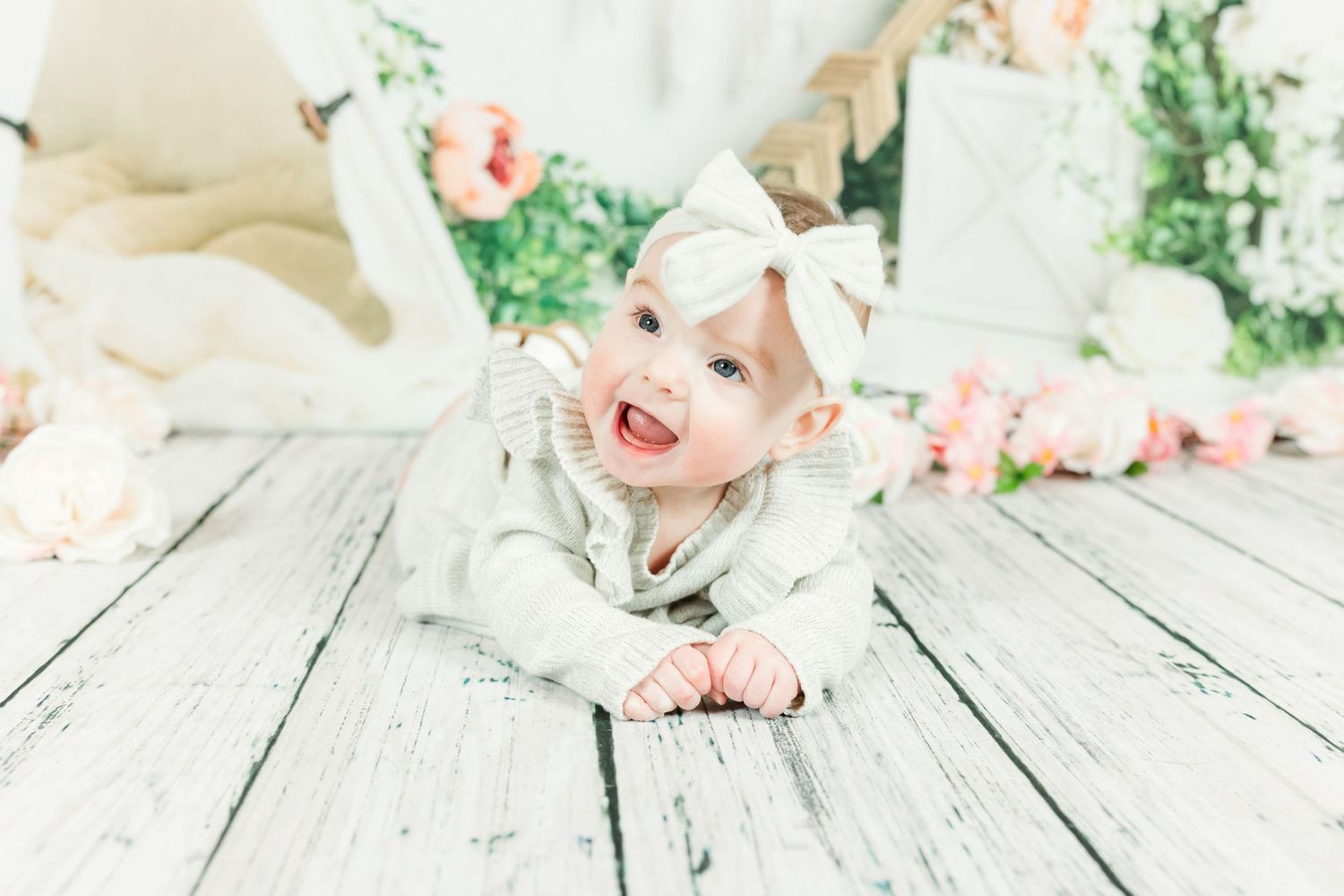 Baby in white outfit lies on wooden floor with floral backdrop during photo session.