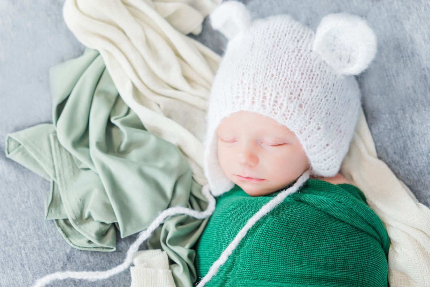 Sleeping infant wearing white knit hat against soft gray and mint green fabrics.
