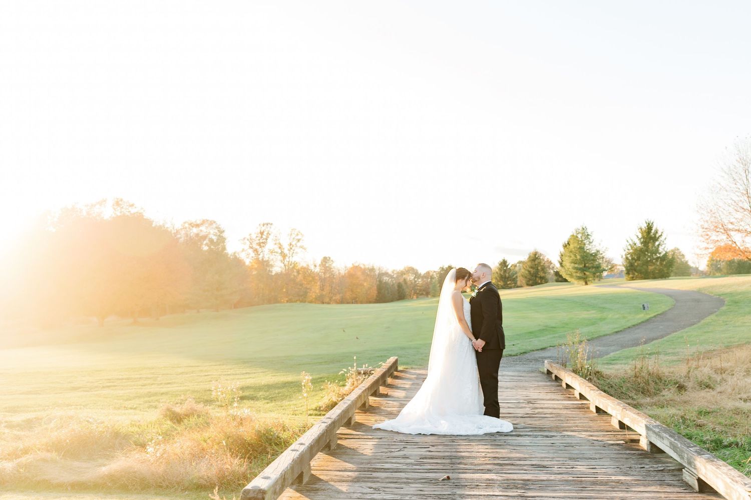 Bride and groom share a romantic moment on a wooden bridge at golden hour with lush green fields in the background.