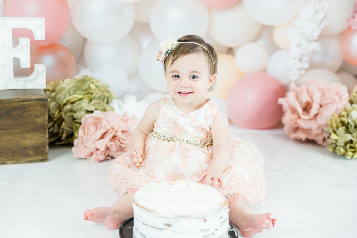 Baby in pink tutu dress sits next to birthday cake during first birthday photoshoot.
