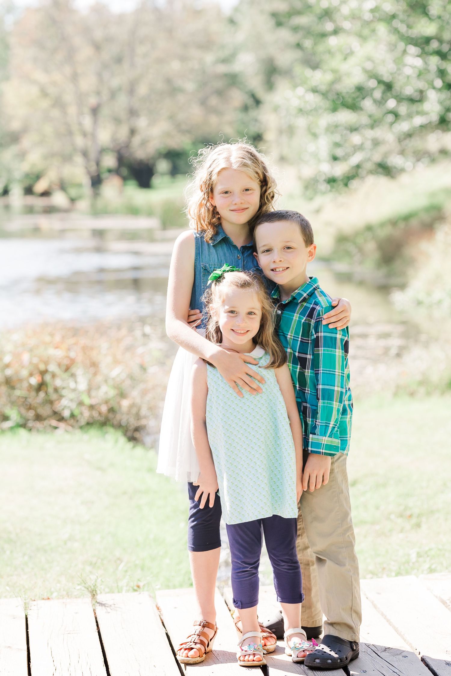 Three children stand together on a wooden deck near a peaceful pond.
