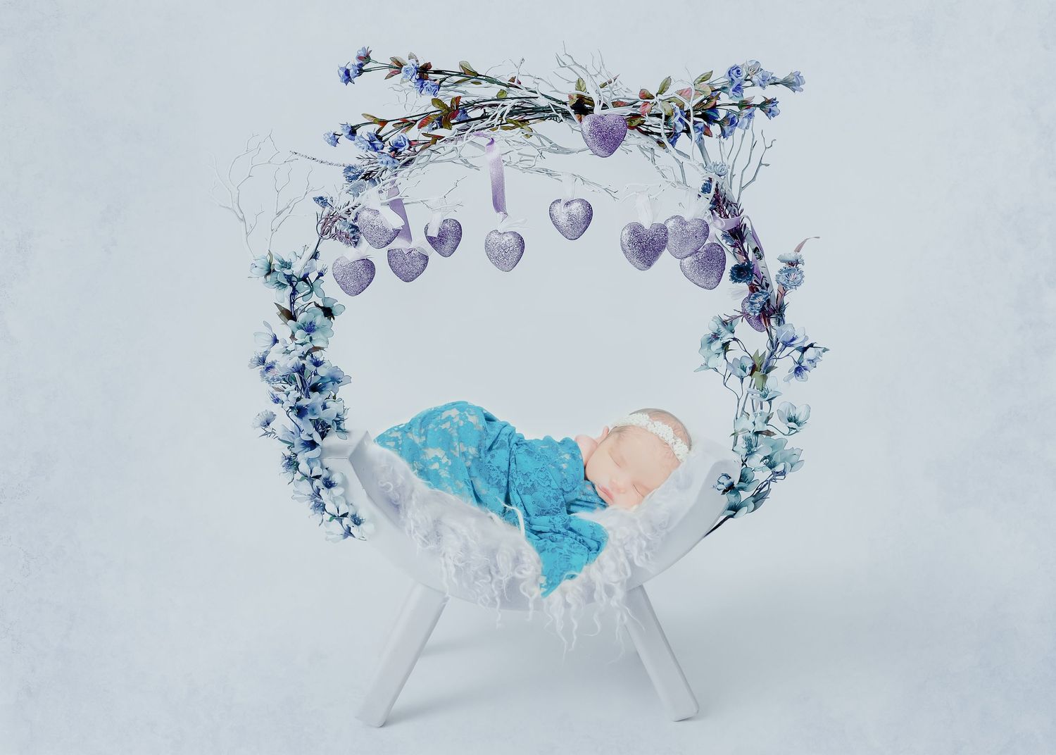 Artistic newborn setup with floral arch and purple hearts against light blue background.