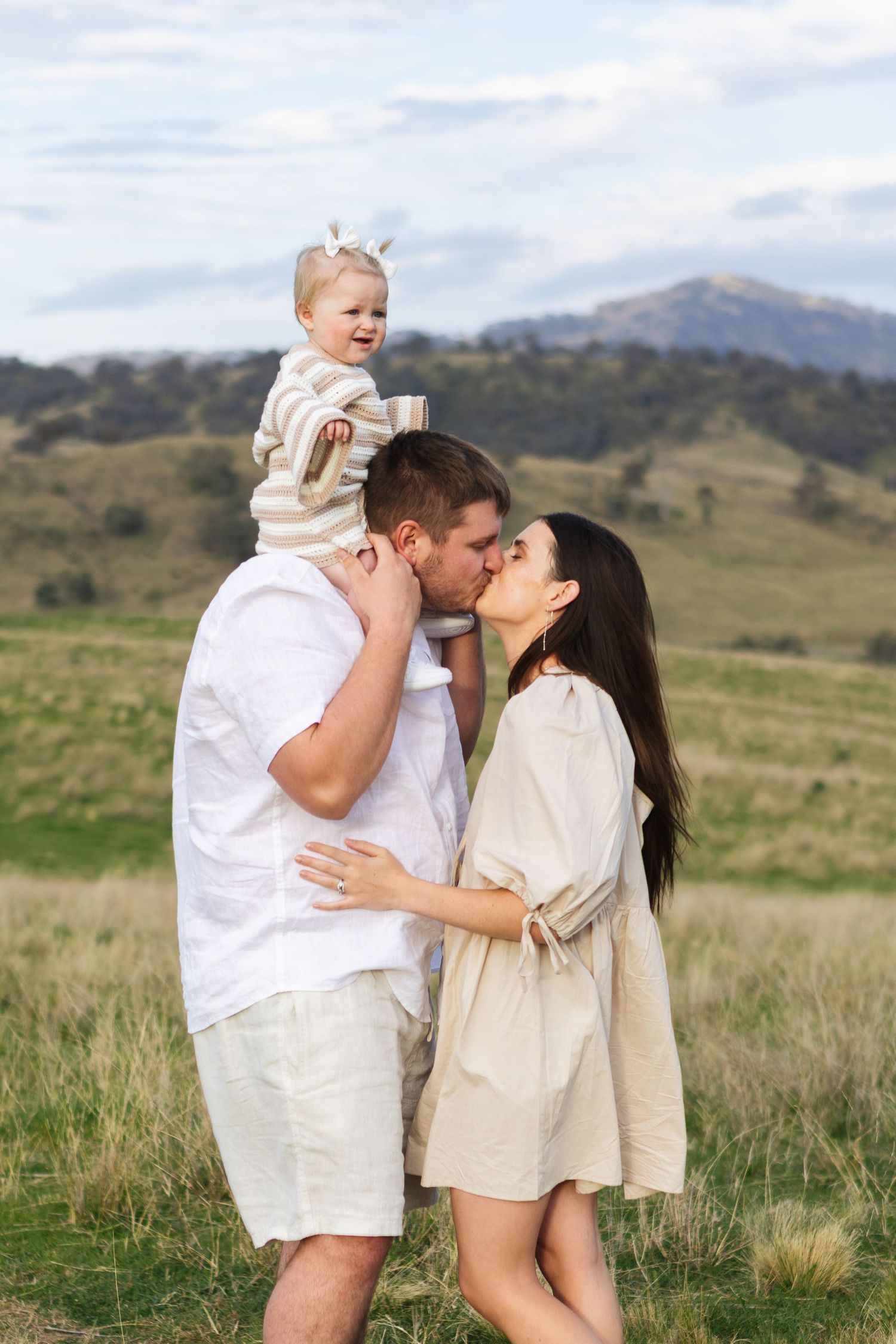mother and father kissing with baby on father's shoulders in country paddock at Scone Family Photoshoot