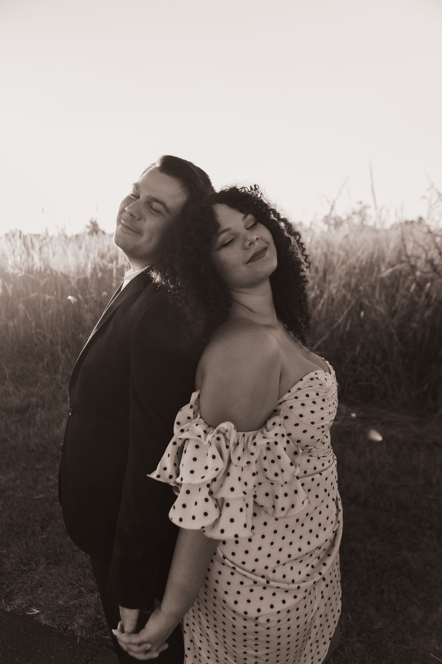 A black and white photo of a newly engaged couple, posing for their engagement photos with VICCI PHOTO, standing back to back in a field