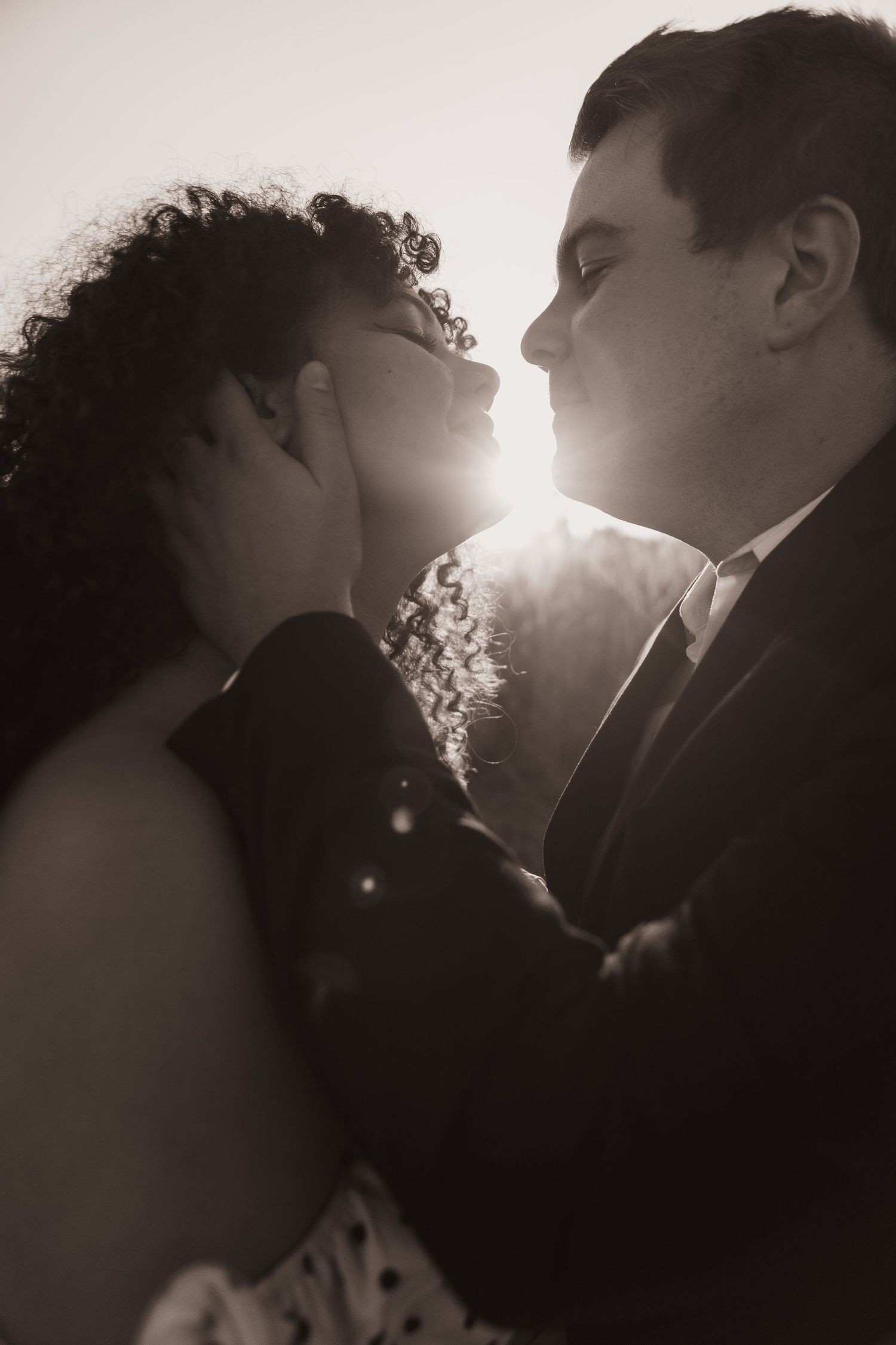 A black and white photo of a newly engaged couple, about to embrace in a kiss with the sunset shining through between them.