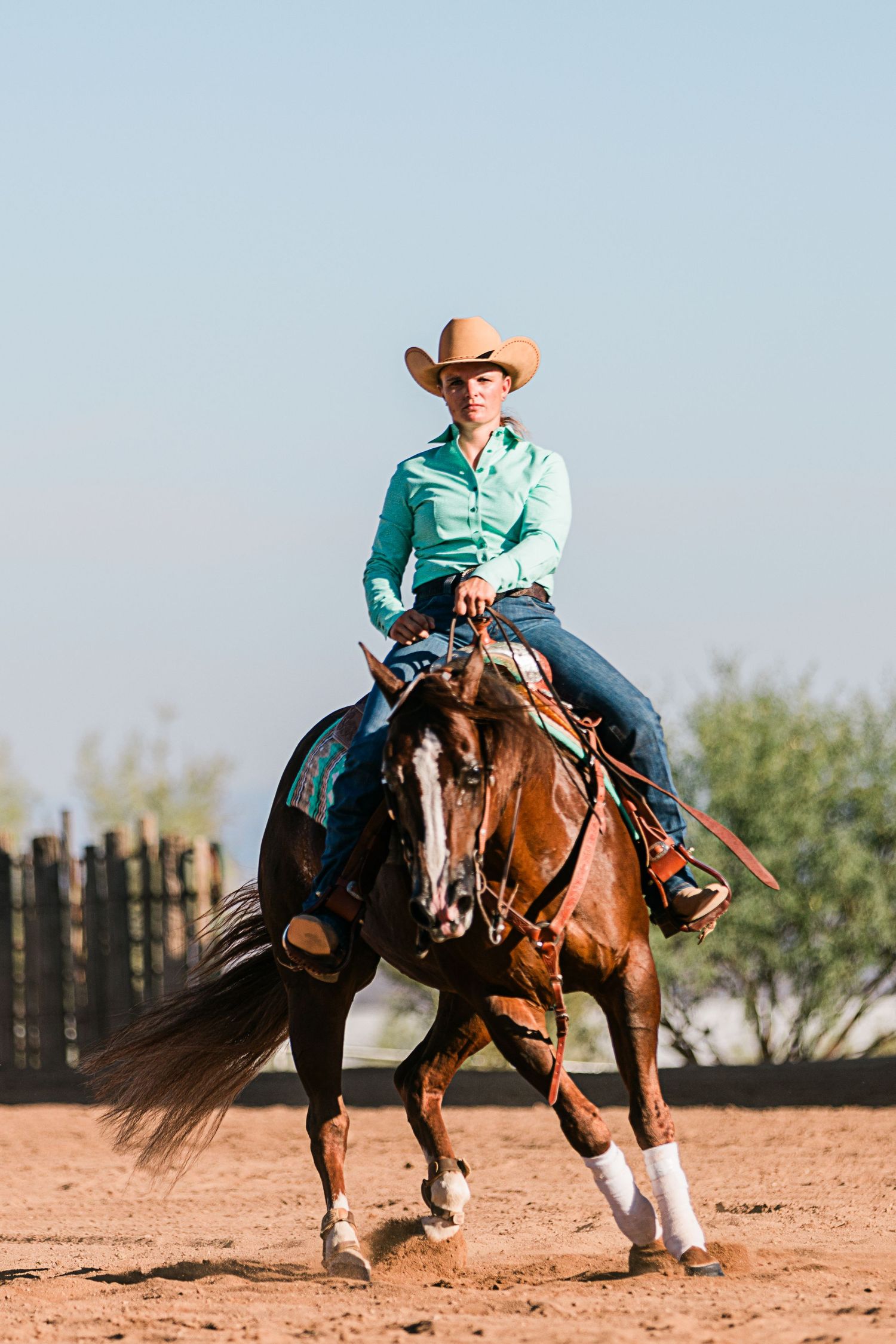 Western rider in turquoise shirt and cowboy hat riding brown horse in sandy arena during equestrian training.