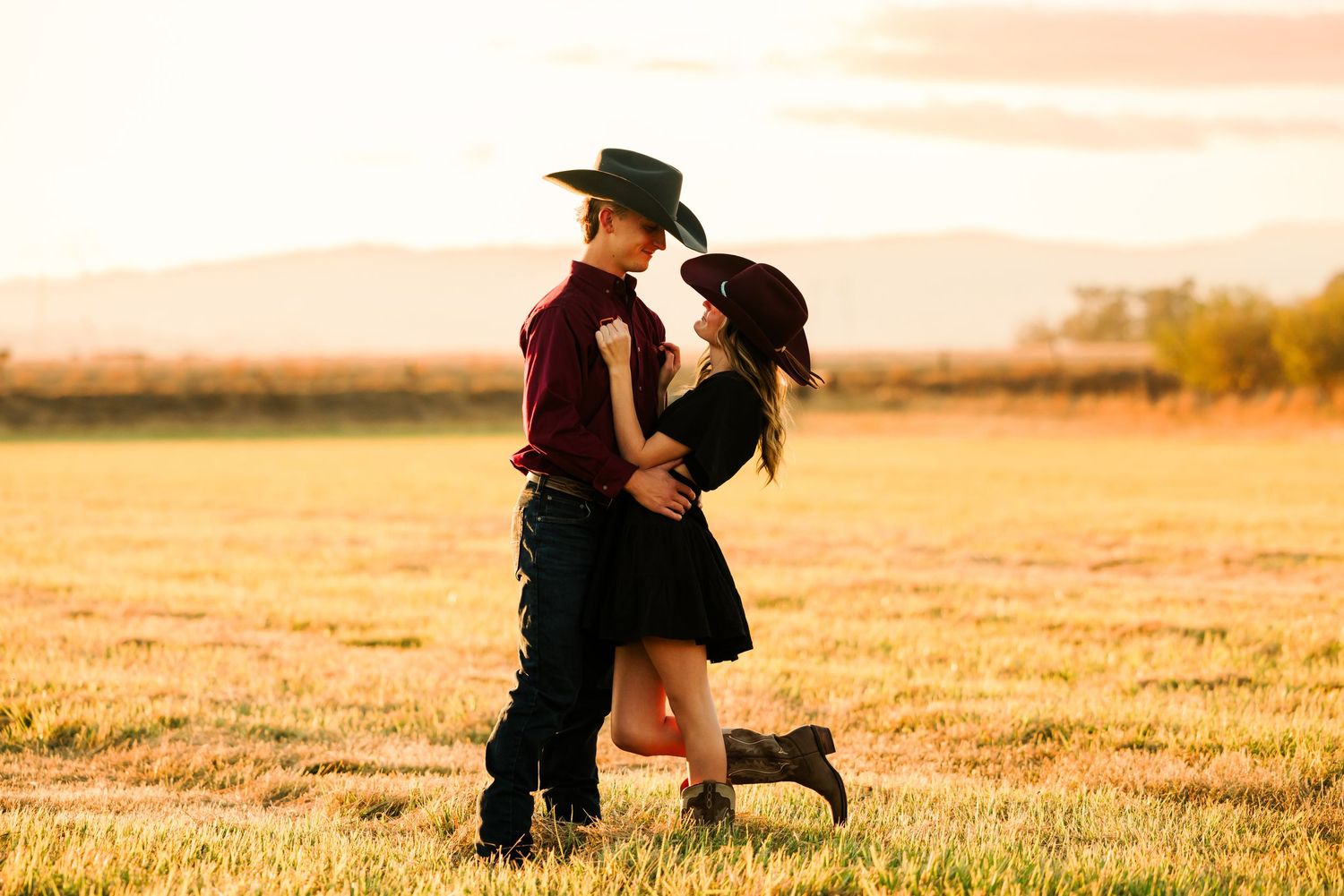A romantic couple embraces in a golden field at sunset, with one wearing a cowboy hat and the other in a black dress.