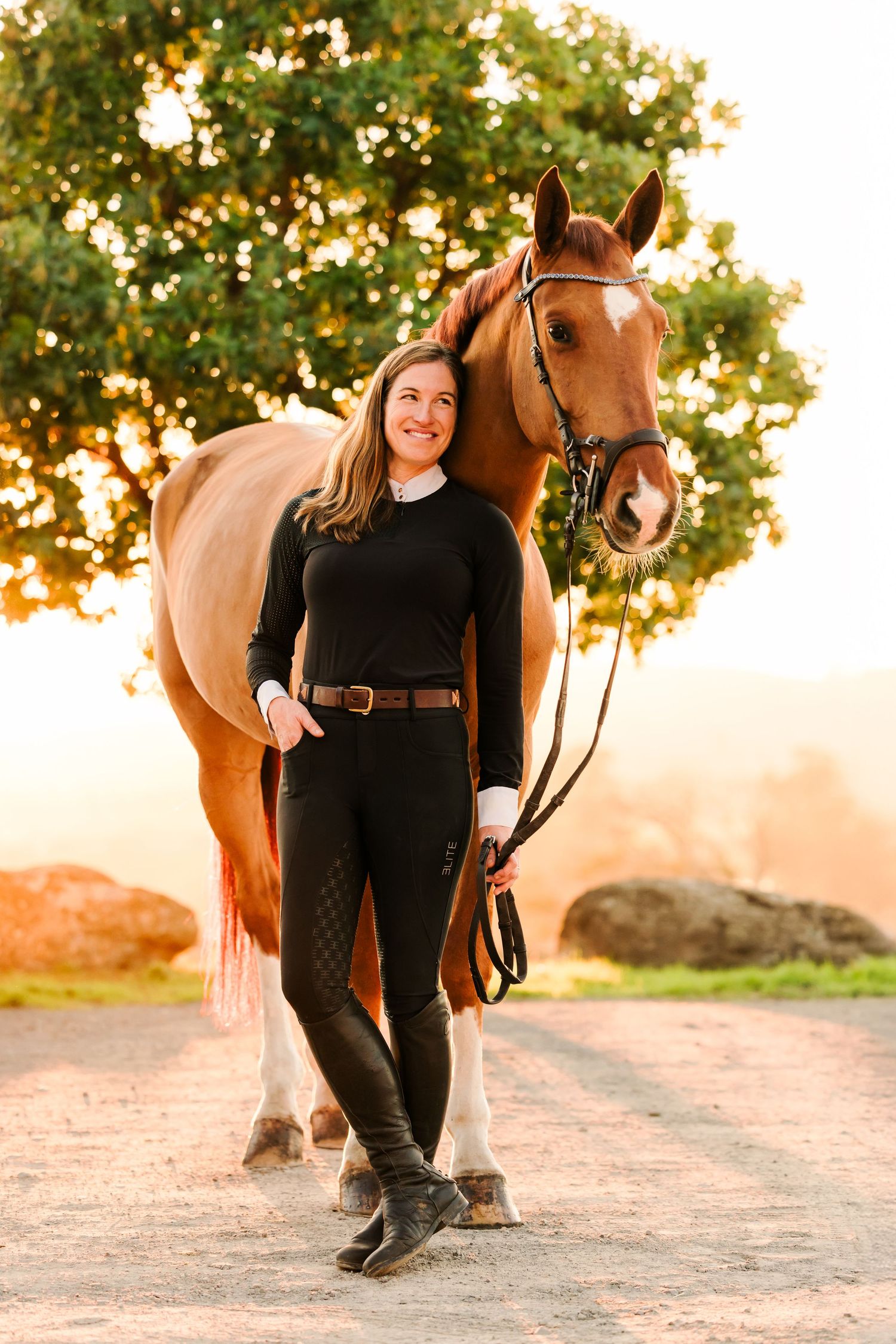 A woman in black clothing poses with a brown horse on a dirt path during golden hour sunset.