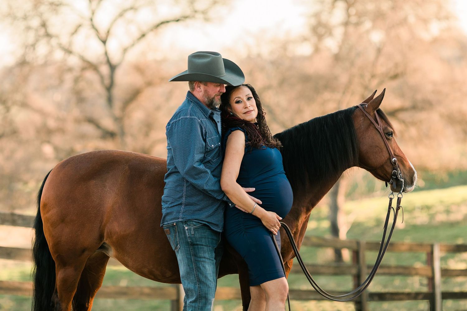 A couple poses with their horse during a romantic engagement photoshoot in a rustic outdoor setting at sunset.