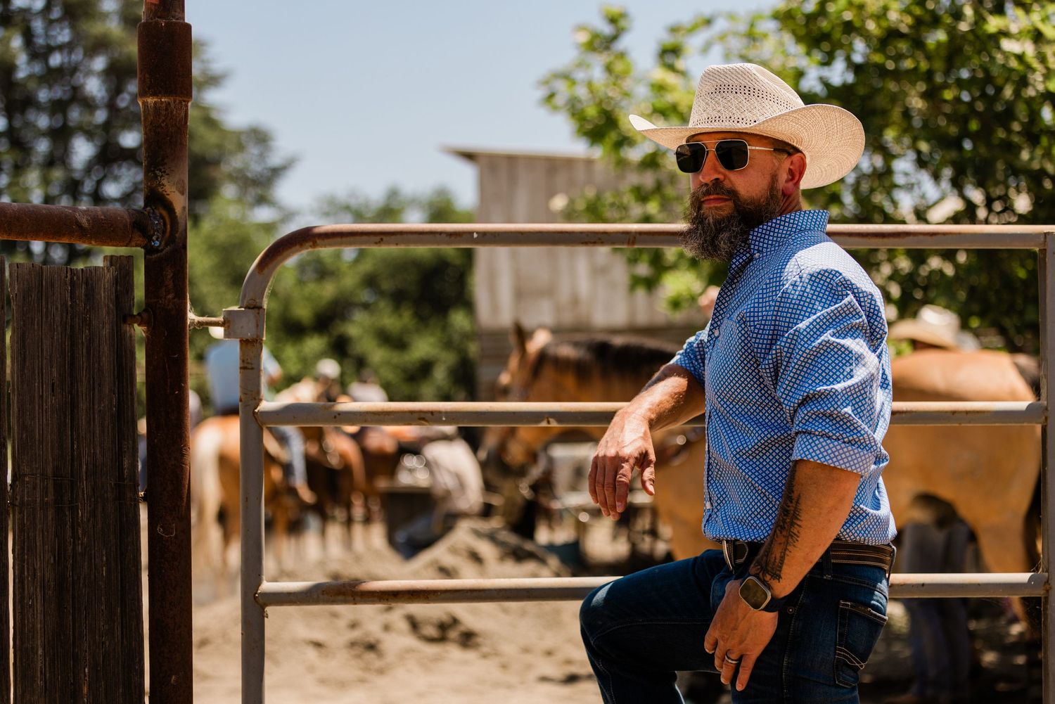 Cowboy in denim and hat standing by ranch fence with cattle in background.