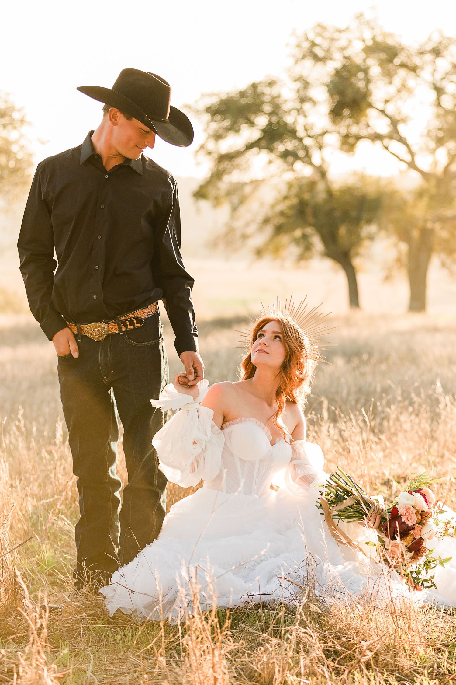 A romantic western-themed couple in black and white attire pose in a golden field at sunset.
