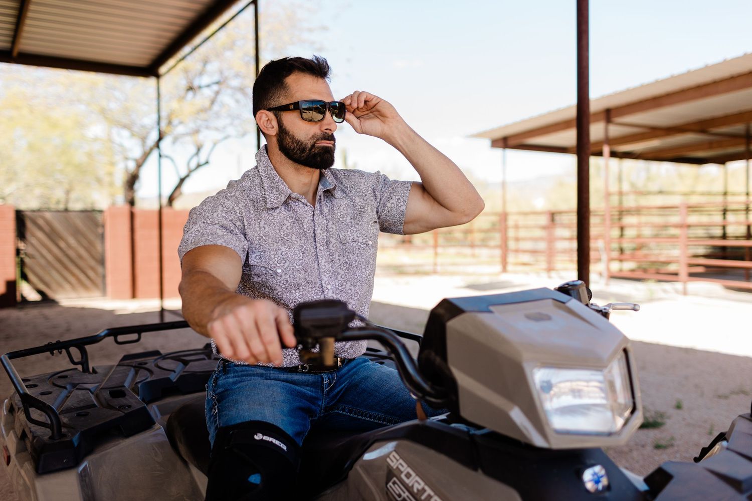 ATV rider adjusting sunglasses while seated on vehicle under covered structure.