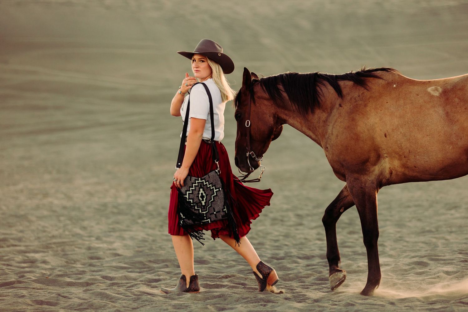 A person in cowboy attire walks along a beach with a brown horse in an atmospheric setting.