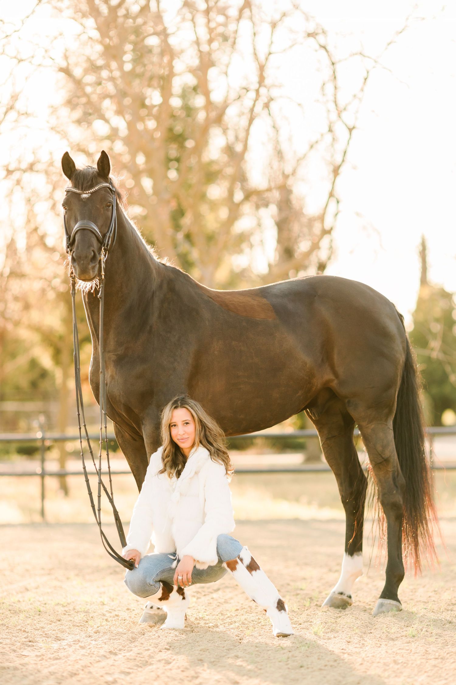A black horse stands beside someone in jeans and white sneakers during a golden sunset photo session.