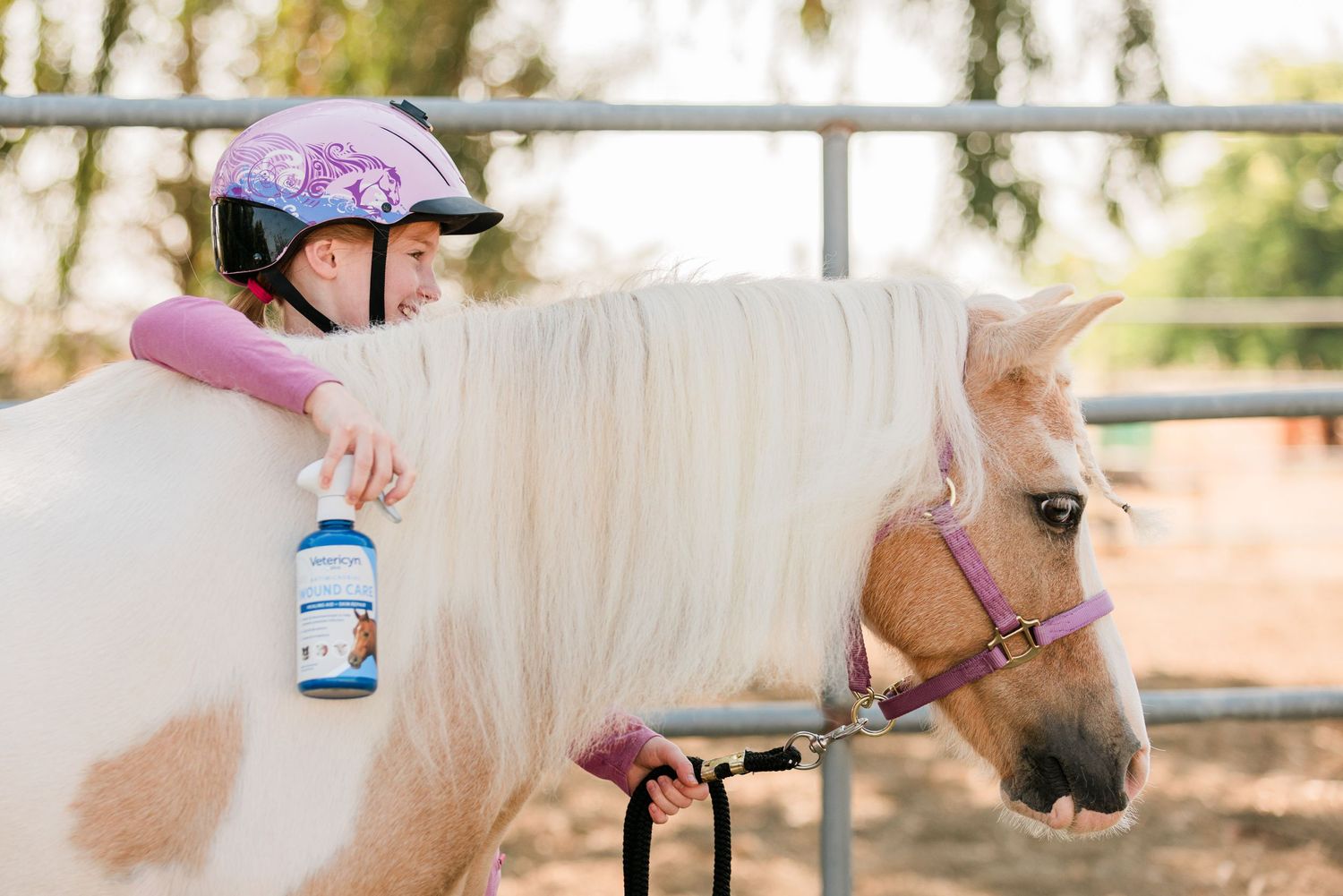 Young rider wearing pink helmet leaning on white pony with blue water bottle.
