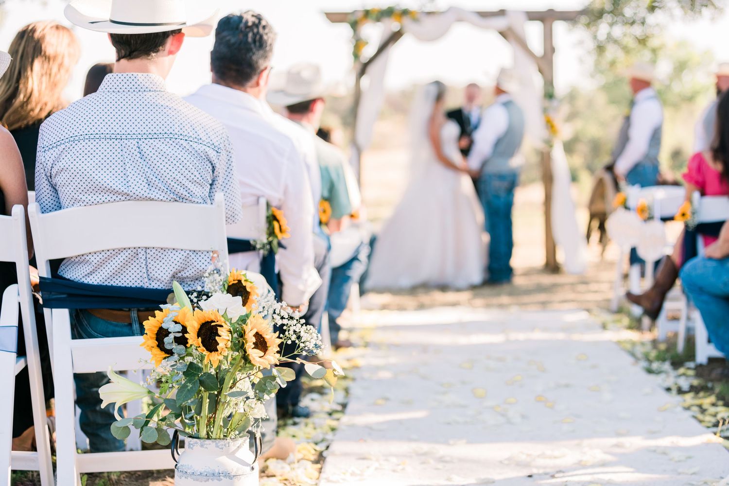 Wedding guests sit in white chairs outdoors during an intimate garden ceremony with bright yellow floral arrangements.