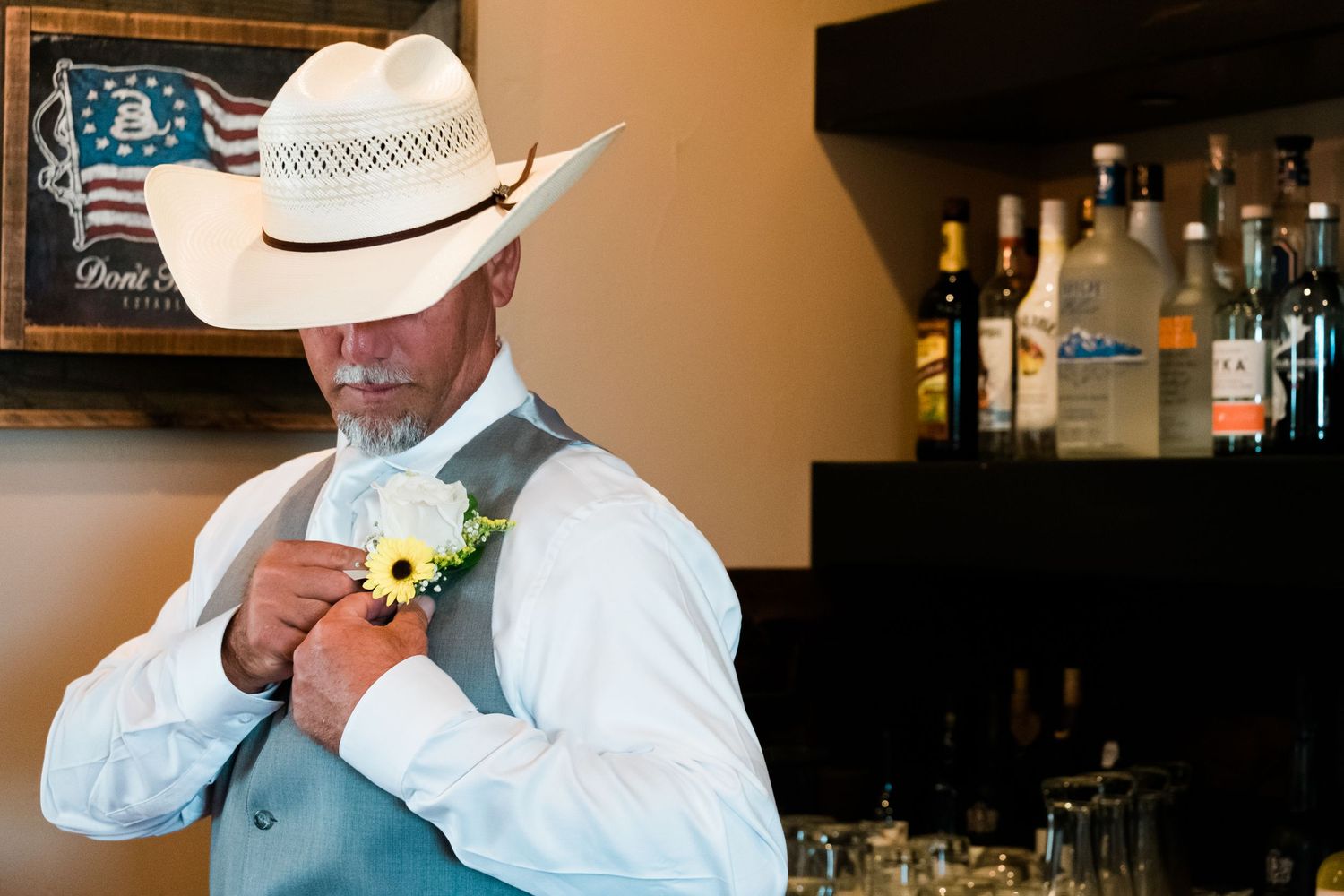 A person in a cowboy hat and white button-down shirt adjusts a yellow floral boutonniere at a bar.