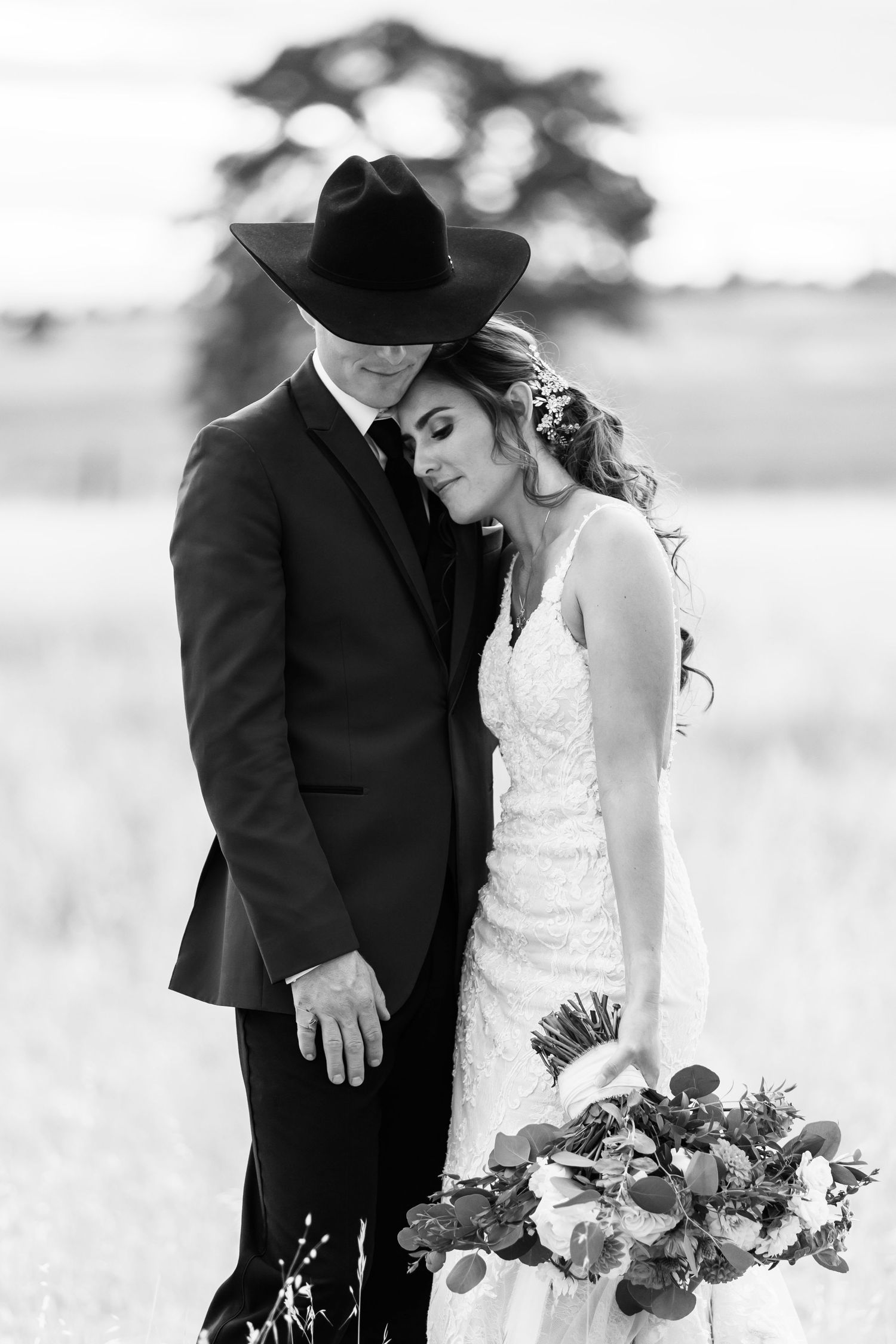 A romantic black and white photo of a bride and groom in western attire standing in a field with a bouquet.