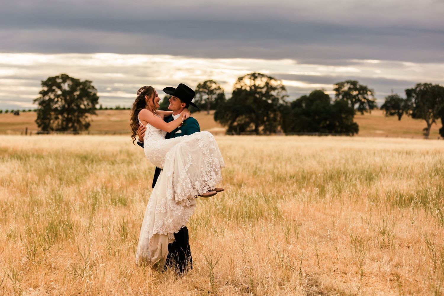 Two people share an intimate embrace in a golden sunlit field of tall grass at sunset.