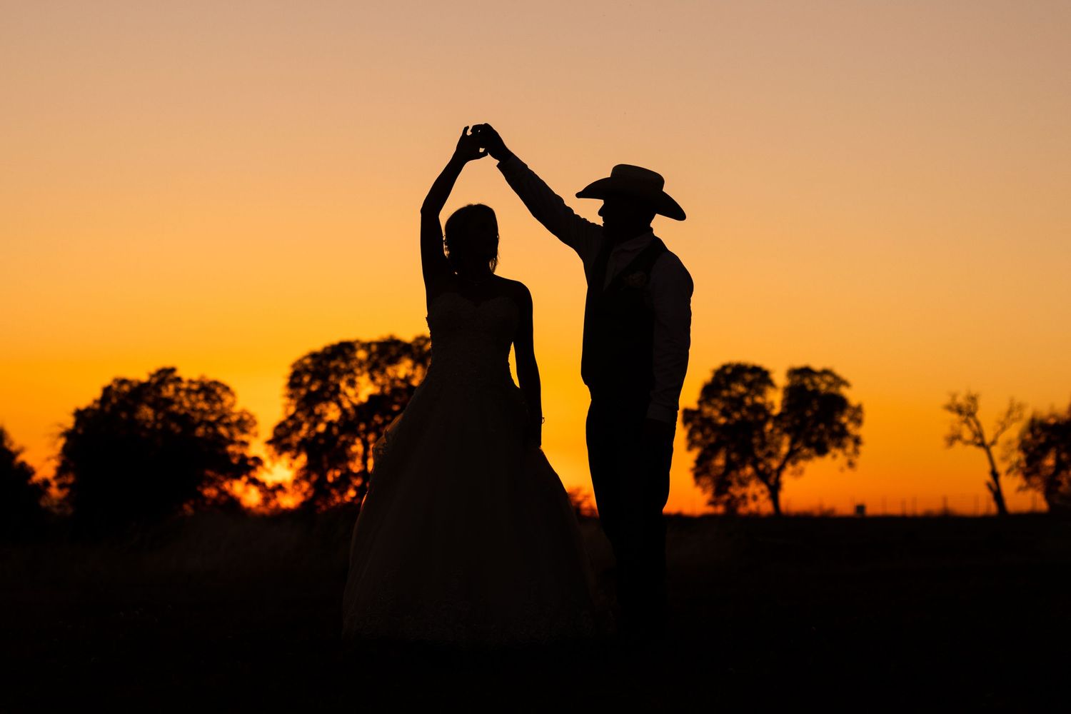 Silhouetted figures stand against vibrant orange sunset sky with trees in the background.
